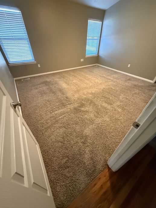 Empty beige-carpeted bedroom with two windows and white trim, viewed from the doorway