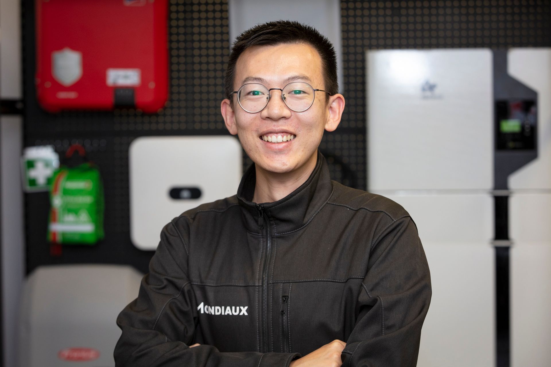 A man wearing glasses and a black jacket is standing in front of a wall of solar panels.