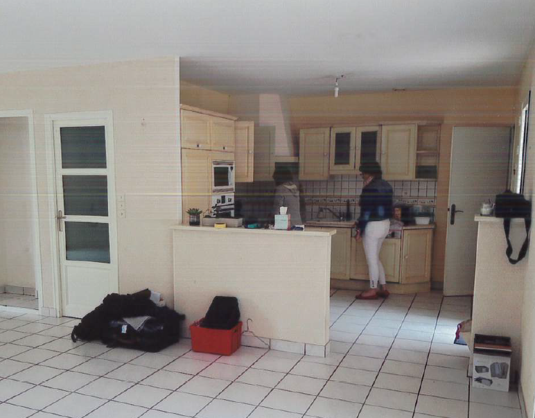 Cuisine avec une personne debout au comptoir ; placards clairs, sol carrelé blanc, espace ouvert.