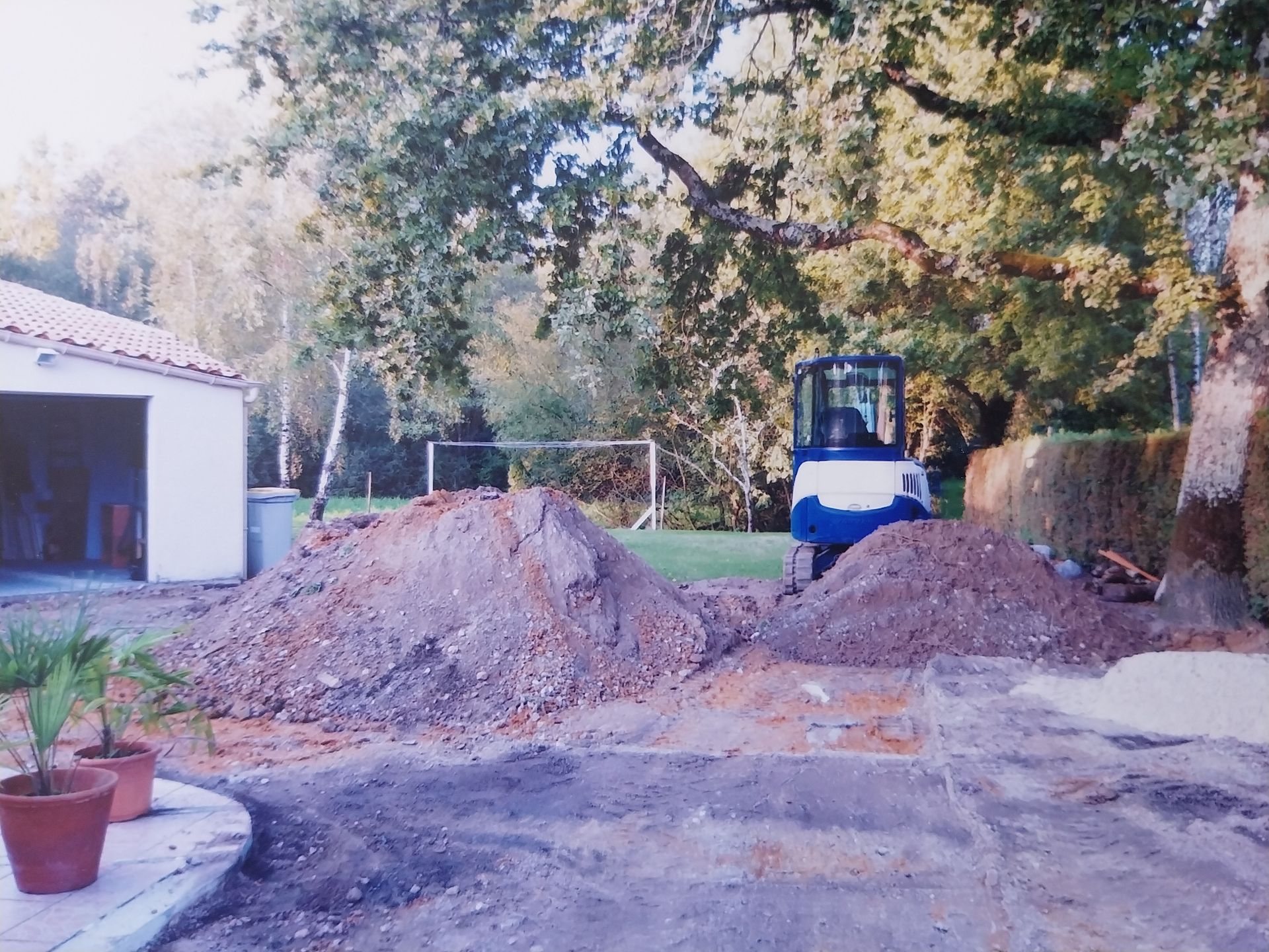 Des tas de terre dans une cour jouxtant un bâtiment blanc abritant un garage, une petite machine et des arbres.