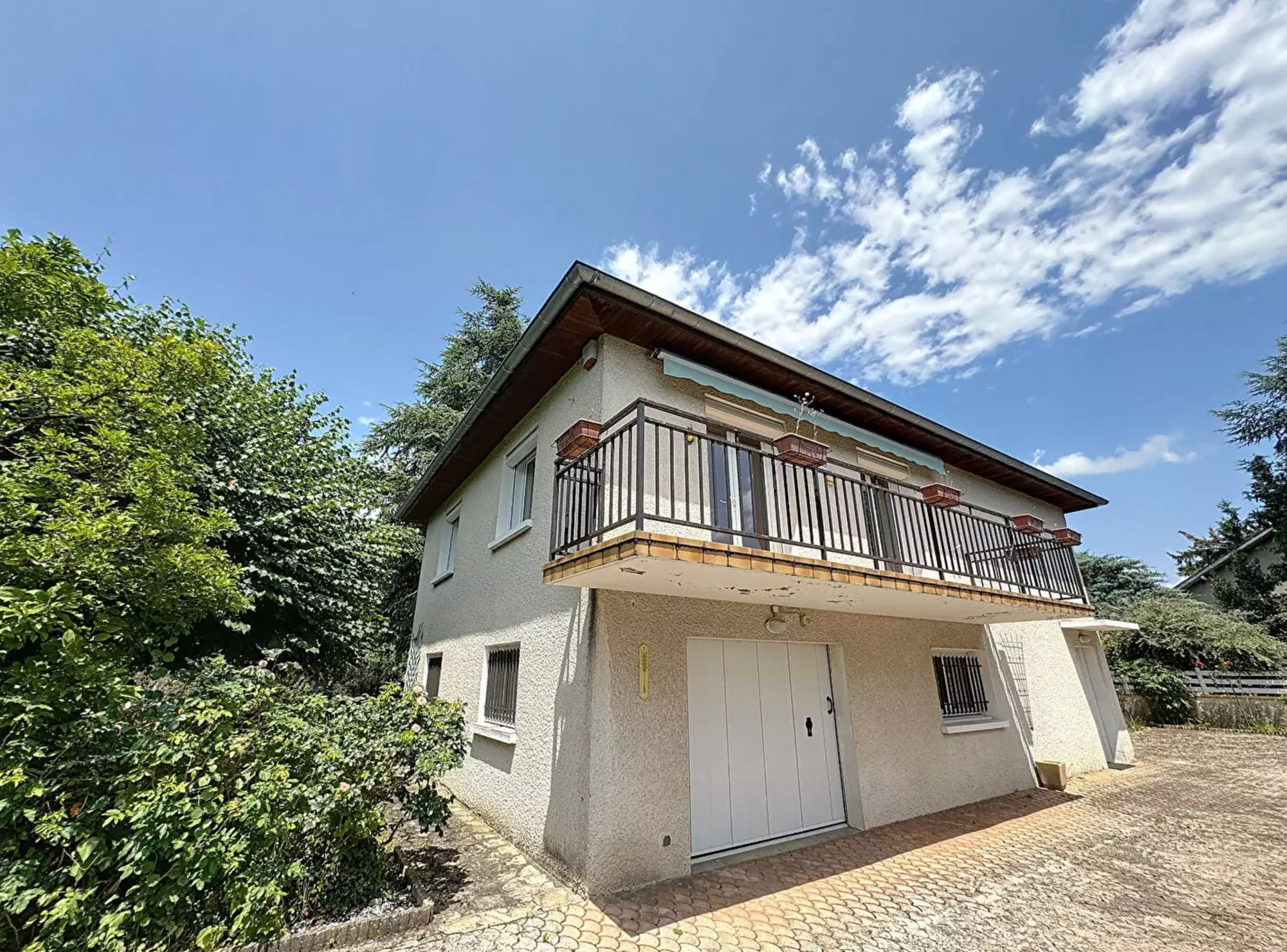 Maison à deux étages avec balcon et portes de garage blanches sous un ciel bleu.