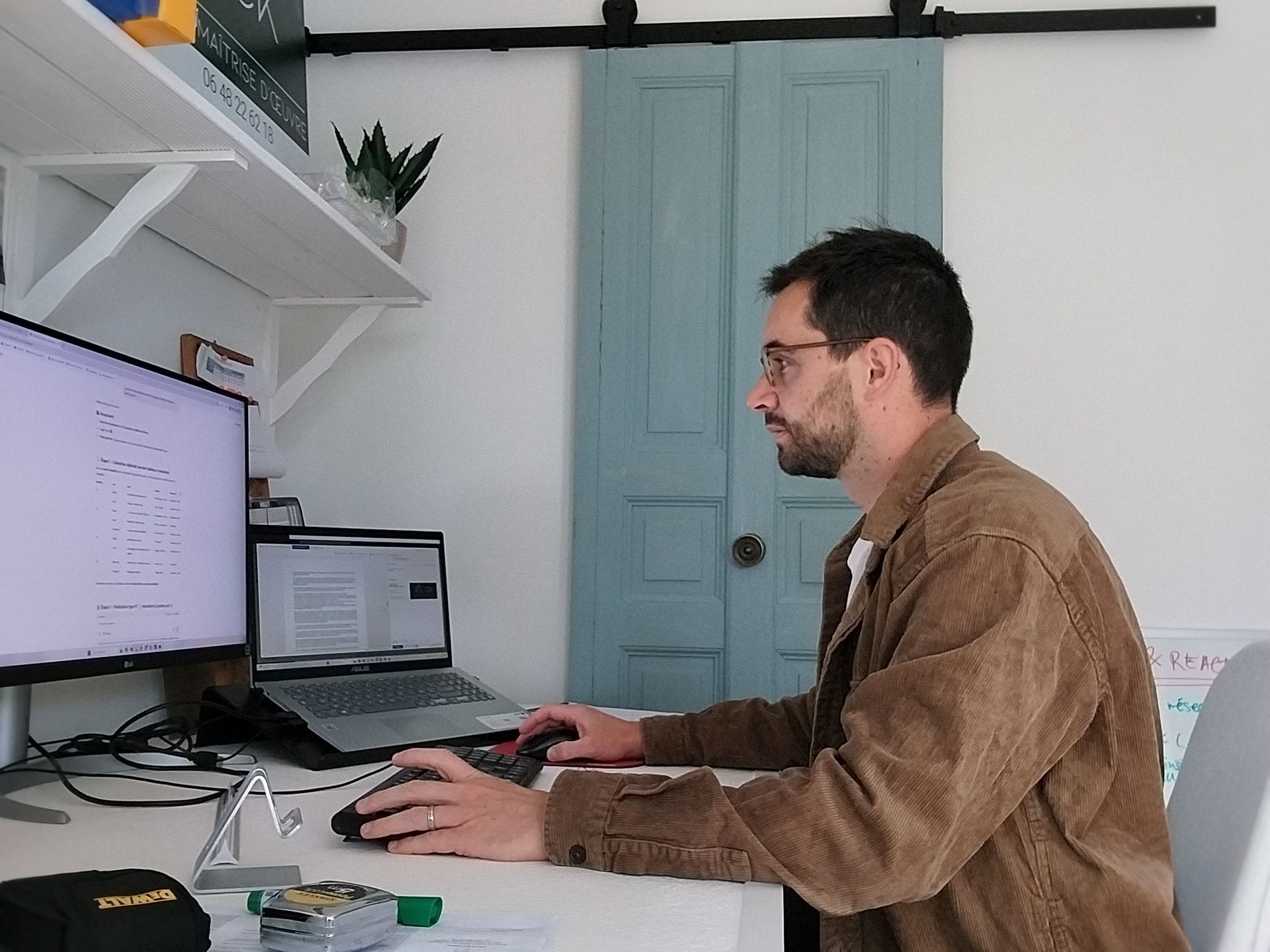 Un homme, assis à son bureau avec deux écrans et un ordinateur portable, portant des lunettes et une veste marron