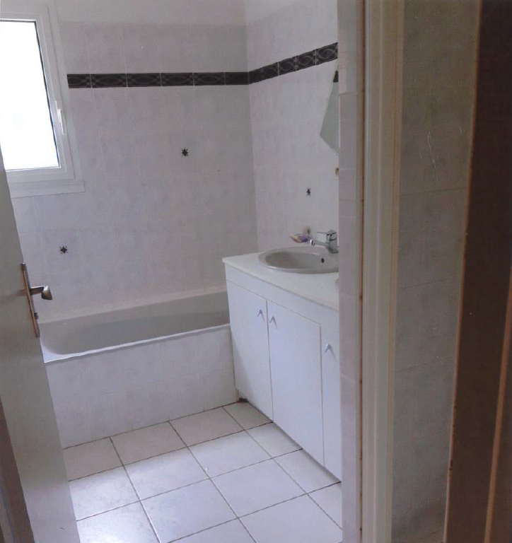 Salle de bains carrelée de blanc, baignoire, lavabo et placards. Une fenêtre se trouve à gauche.