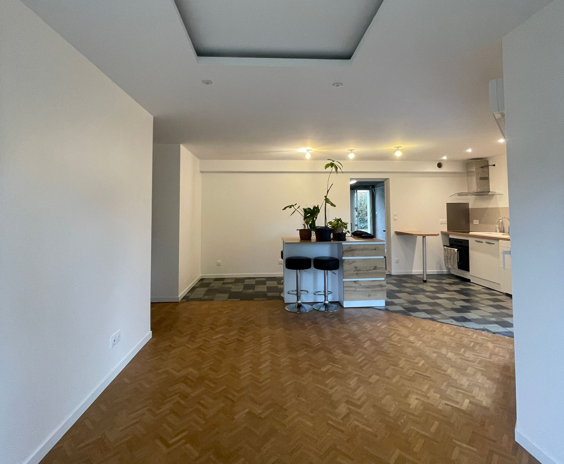 Vue intérieure d'une cuisine et d'un séjour modernes à aire ouverte, avec parquet et îlot central équipé de tabourets de bar.