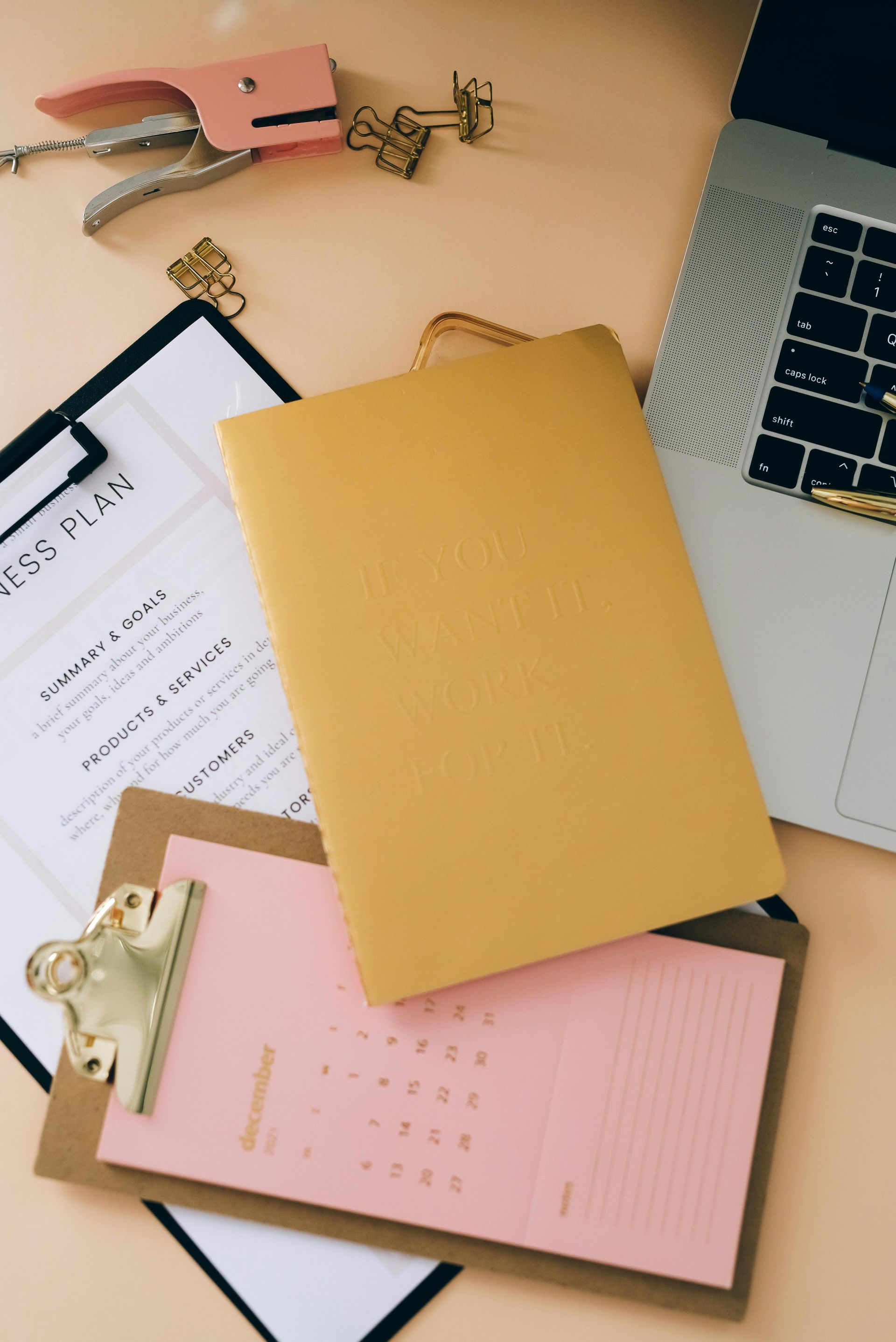 Un cahier jaune sur un calendrier rose et un plan d'affaires sur un bureau avec un ordinateur portable et des fournitures.