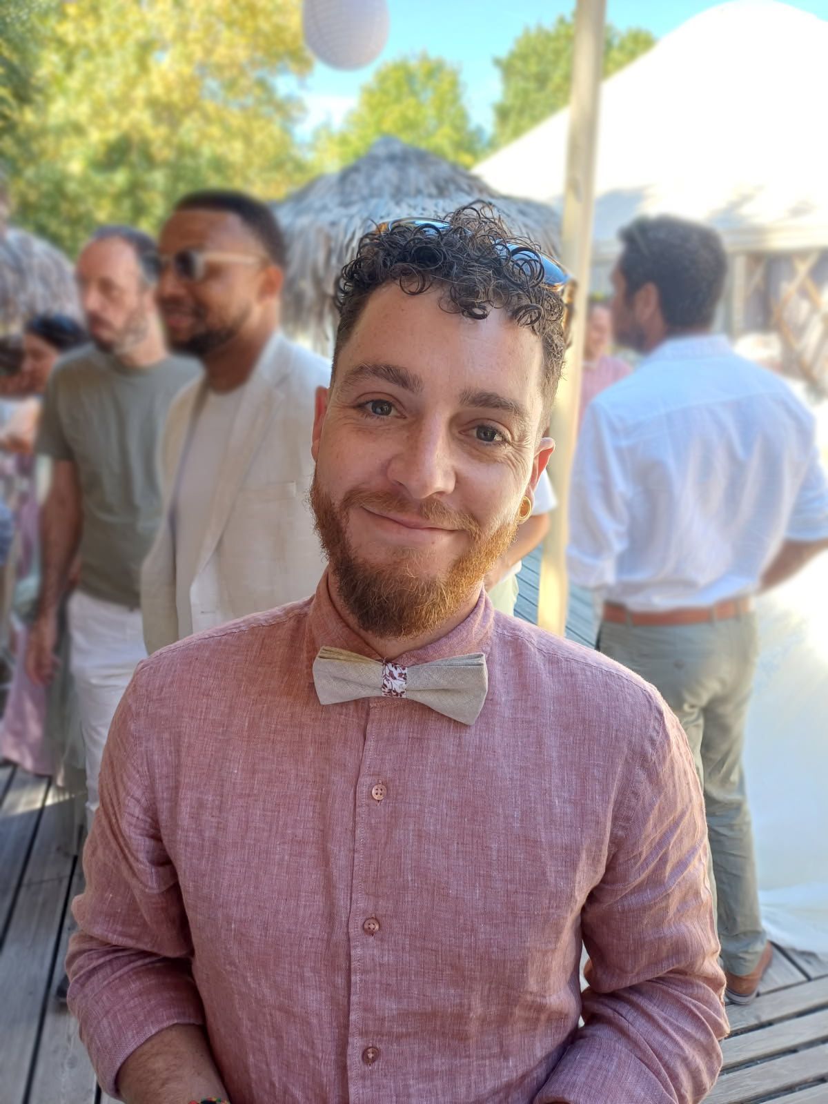 Un homme aux cheveux bouclés et à la barbe souriante, portant une chemise rose et un nœud papillon, se trouve à l'extérieur.