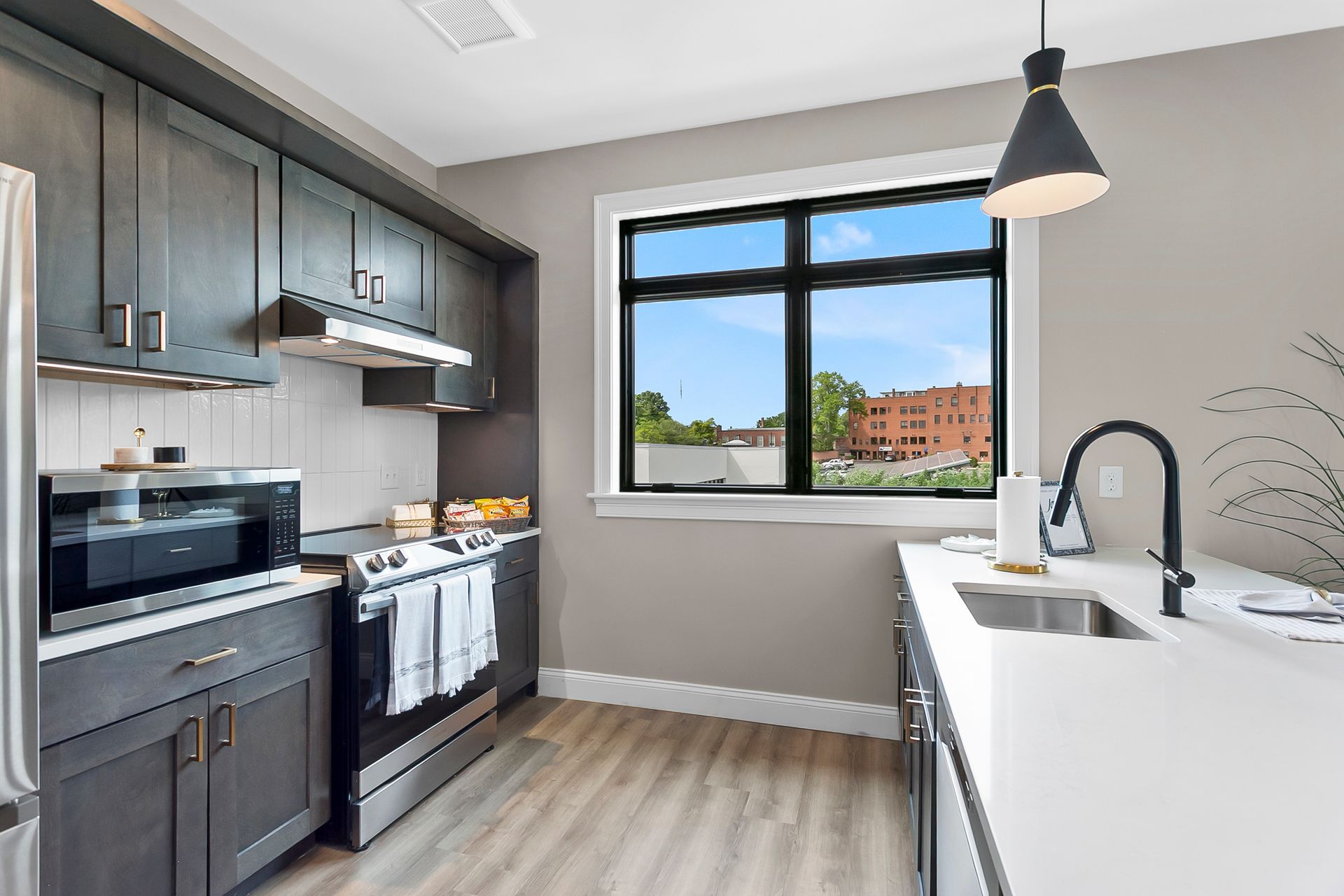 Modern kitchen with dark cabinets, white countertops, pendant light, and a large window overlooking buildings.