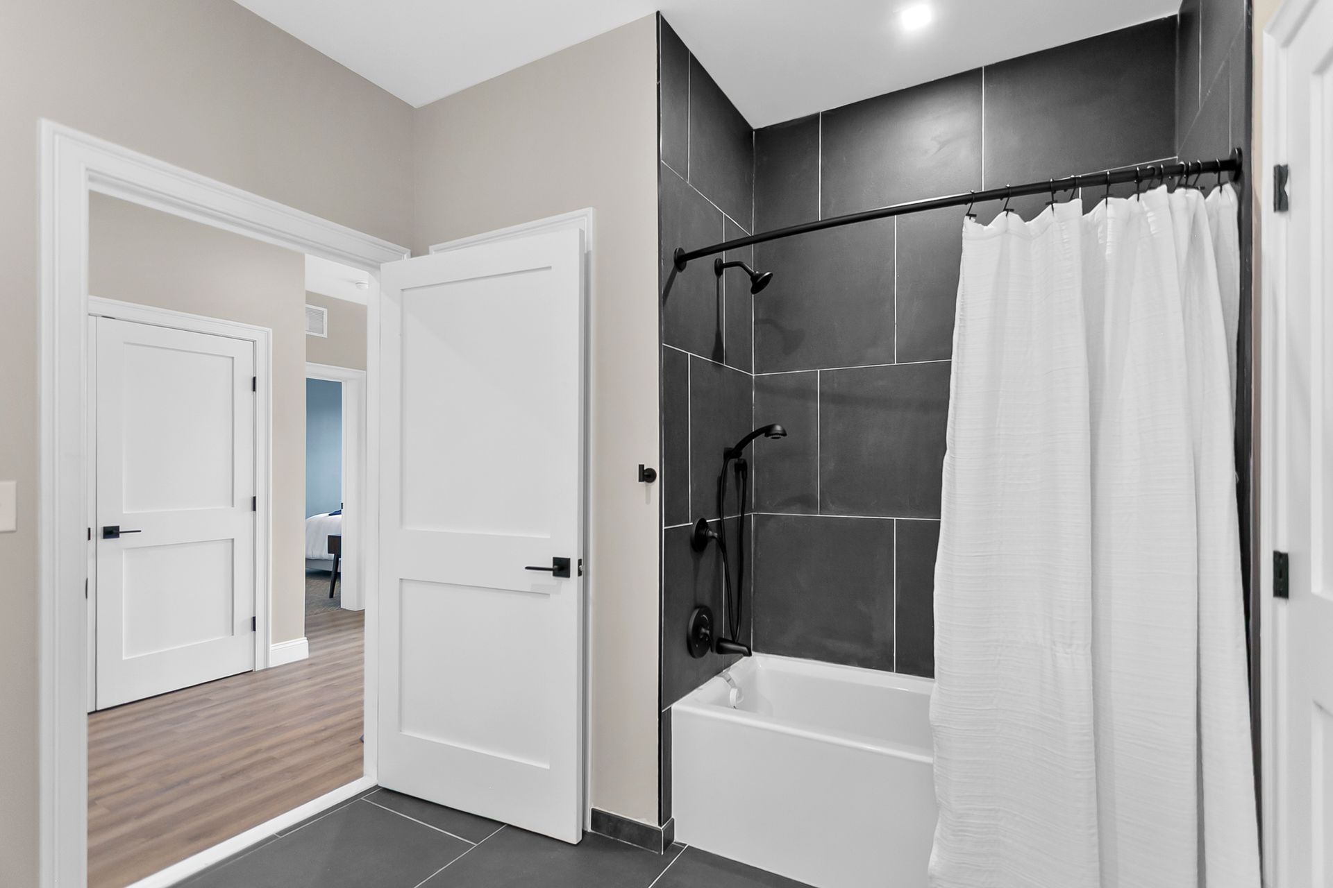 Modern bathroom with dark tile shower, white tub, and open doors to a wood-floored hallway