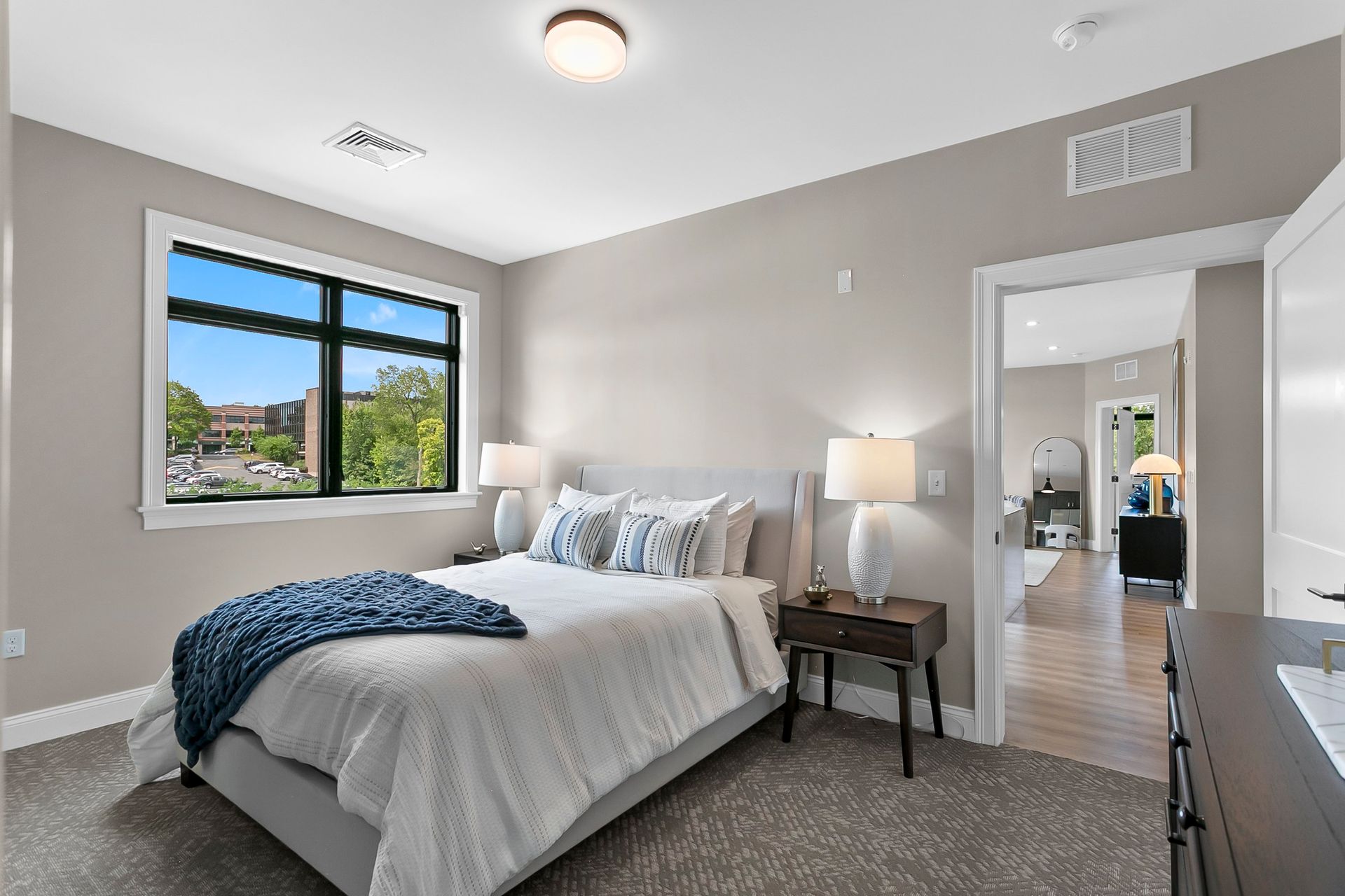 Modern bedroom with gray walls, large window, bed, bedside lamp, and open doorway to a bright hall