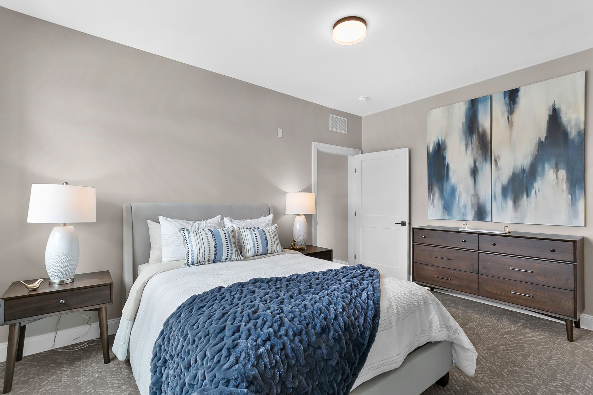 Modern bedroom with a blue quilt, abstract wall art, and a dresser with lamps