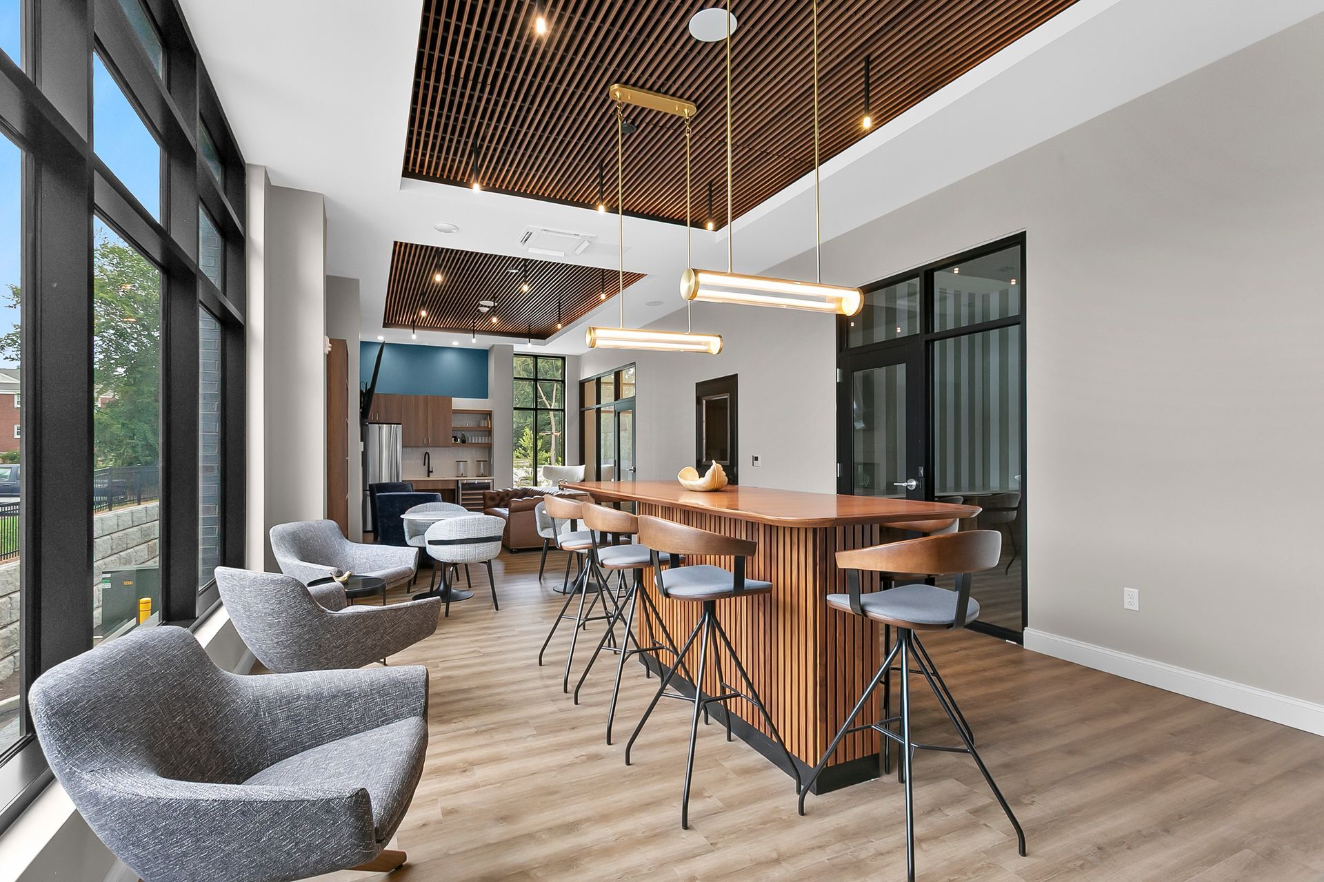Modern lounge with gray chairs, wood tables, and a long communal table under slatted ceiling lights.