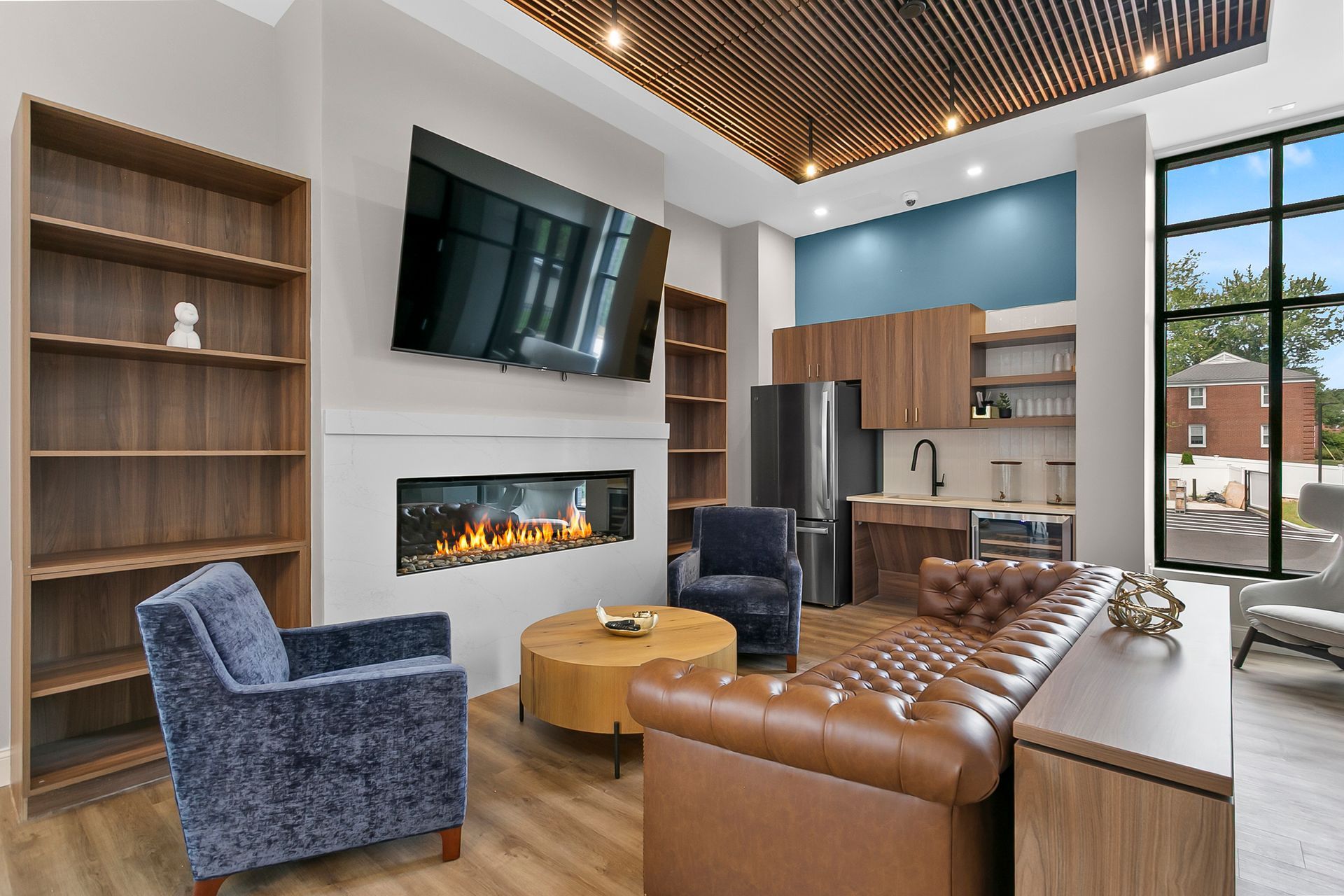 Modern lounge with fireplace, wall TV, blue chairs, tan sofa, and kitchen area with large windows
