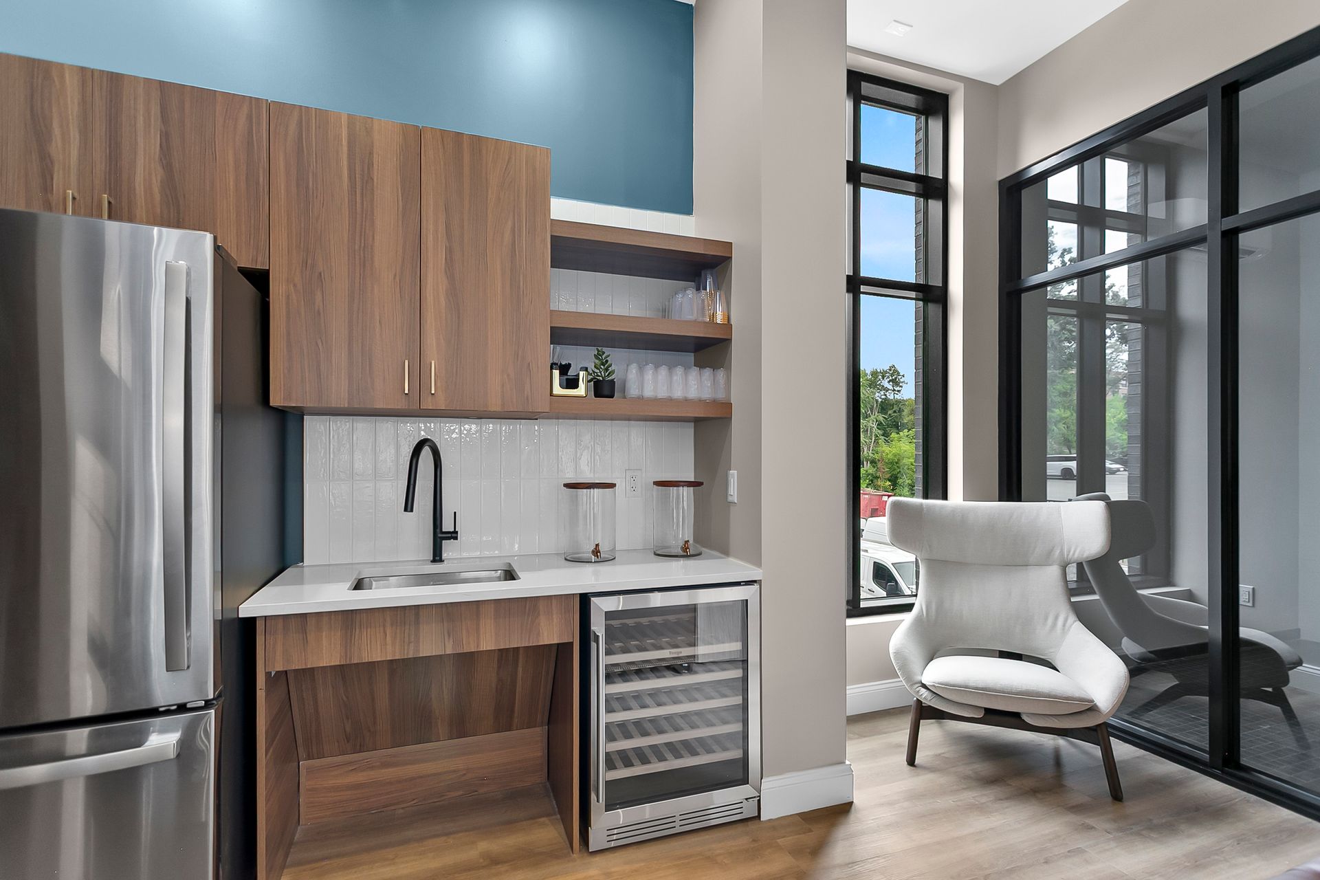 Modern kitchenette with wood cabinets, stainless fridge, wine cooler, and a bright lounge chair by black-framed windows