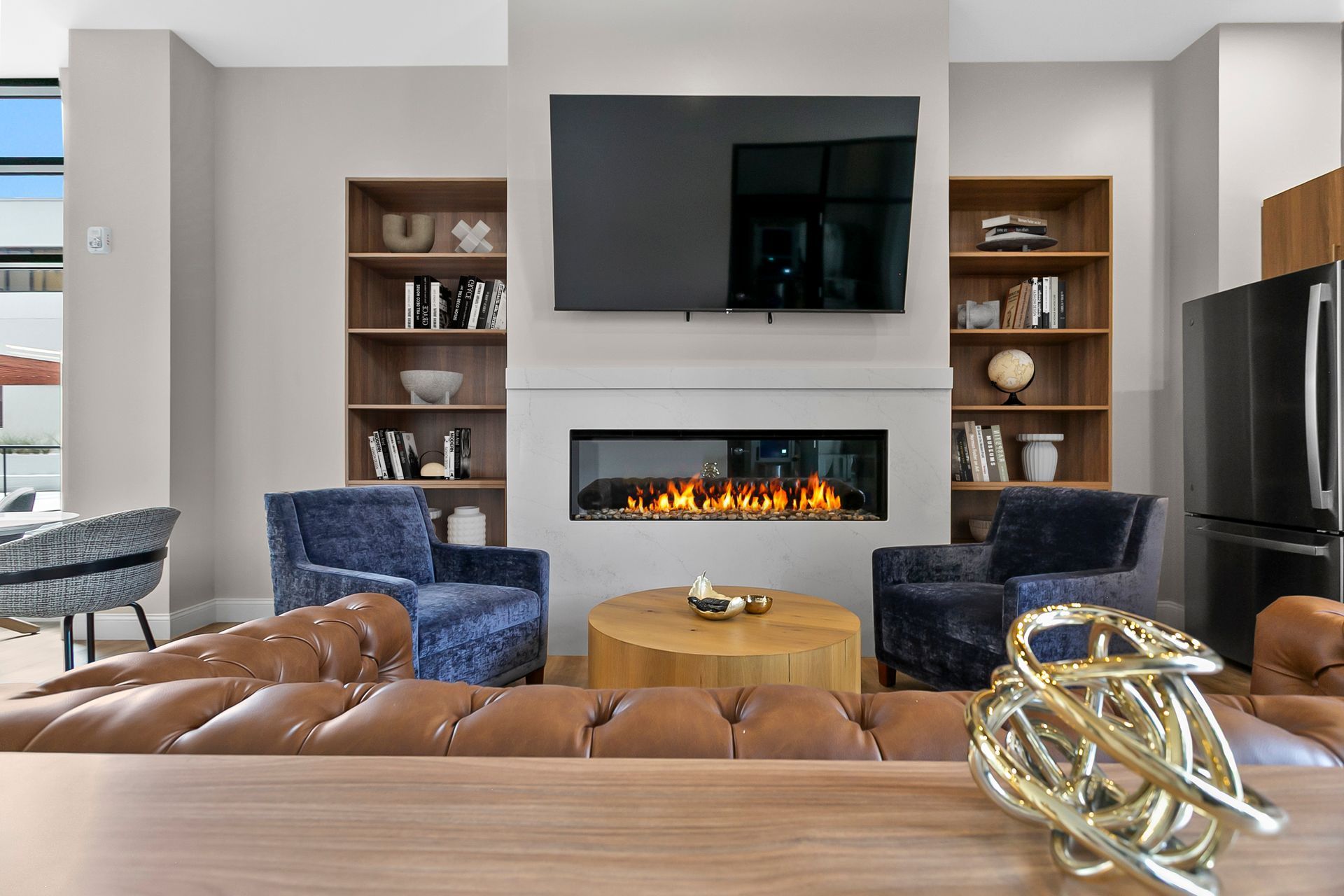 Modern living room with fireplace, wall-mounted TV, blue chairs, tan ottoman, and built-in shelves
