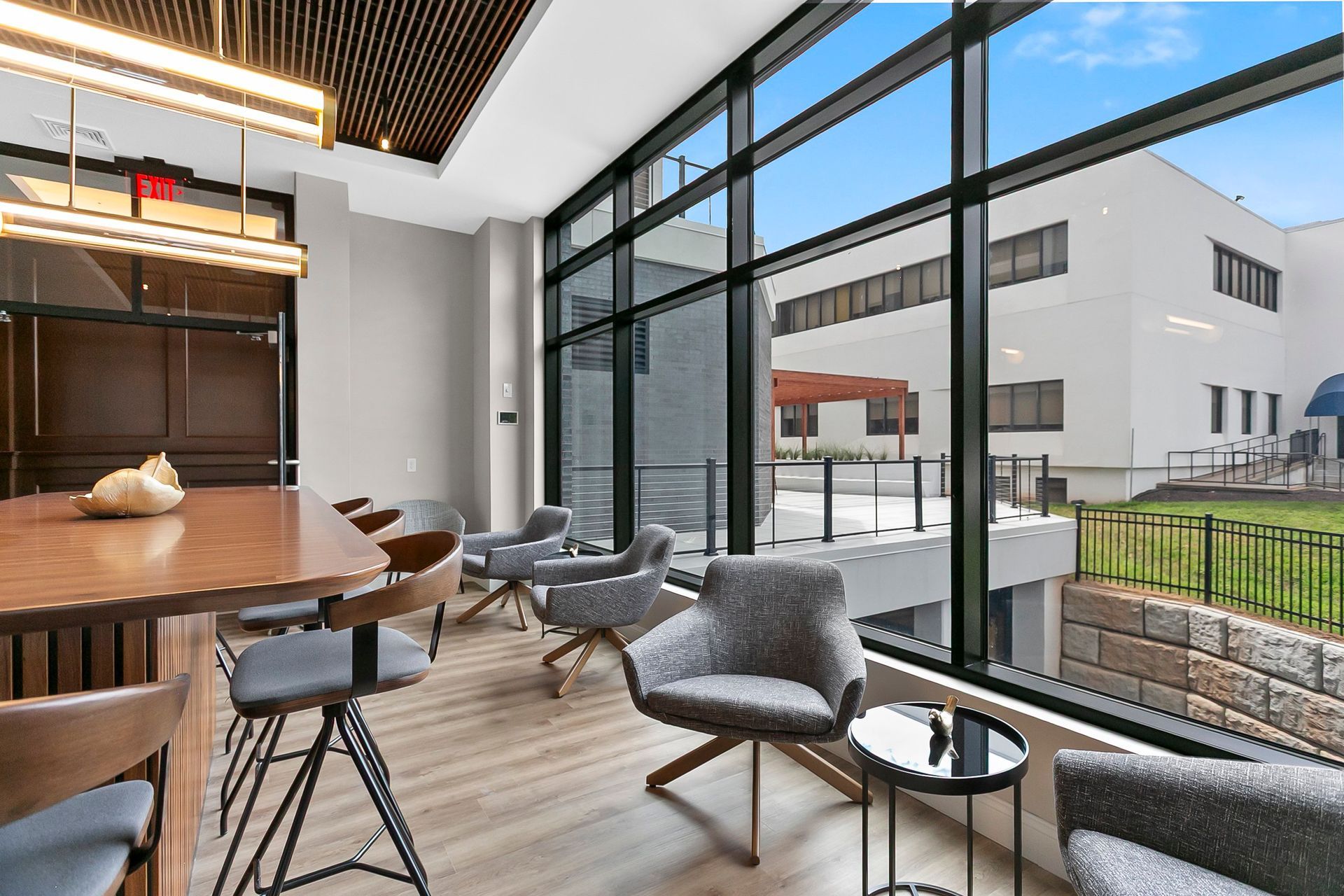 Modern lounge with round wooden table, gray chairs, and floor-to-ceiling windows overlooking a courtyard