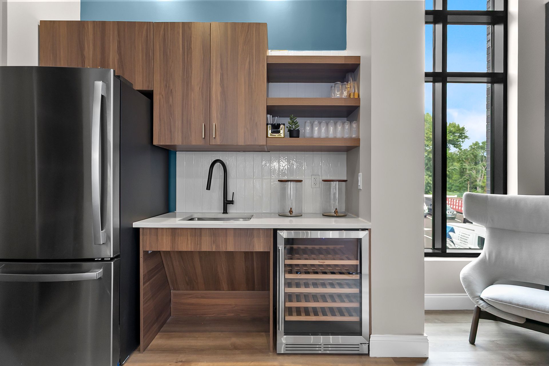 Modern kitchenette with black fridge, wood cabinets, white counter, sink, wine cooler, and large window.