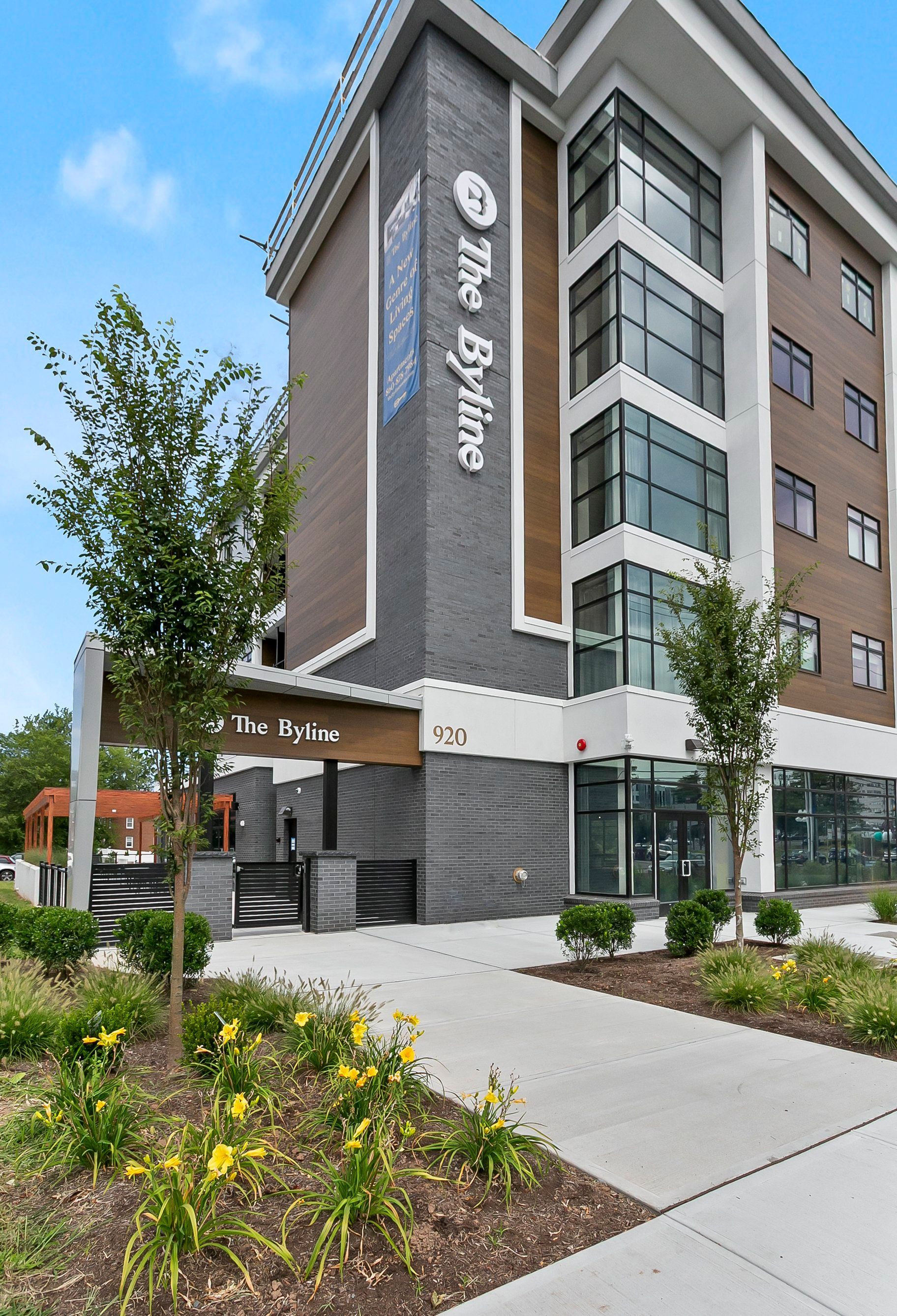 Modern multi-story hotel building with gray brick exterior, glass windows, and landscaped sidewalk entrance