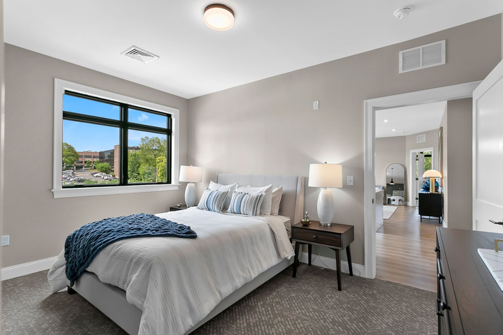 Bright modern bedroom with a bed, bedside lamp, window, and open doorway to a hallway.