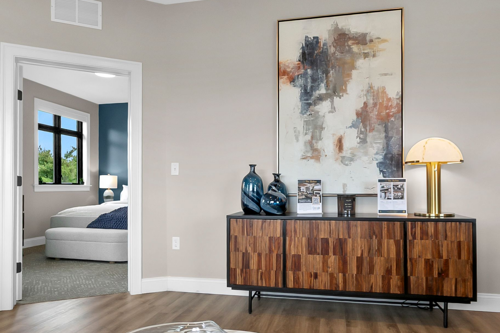 Modern bedroom with a walnut dresser, abstract wall art, and a lit lamp; doorway reveals bed and window.