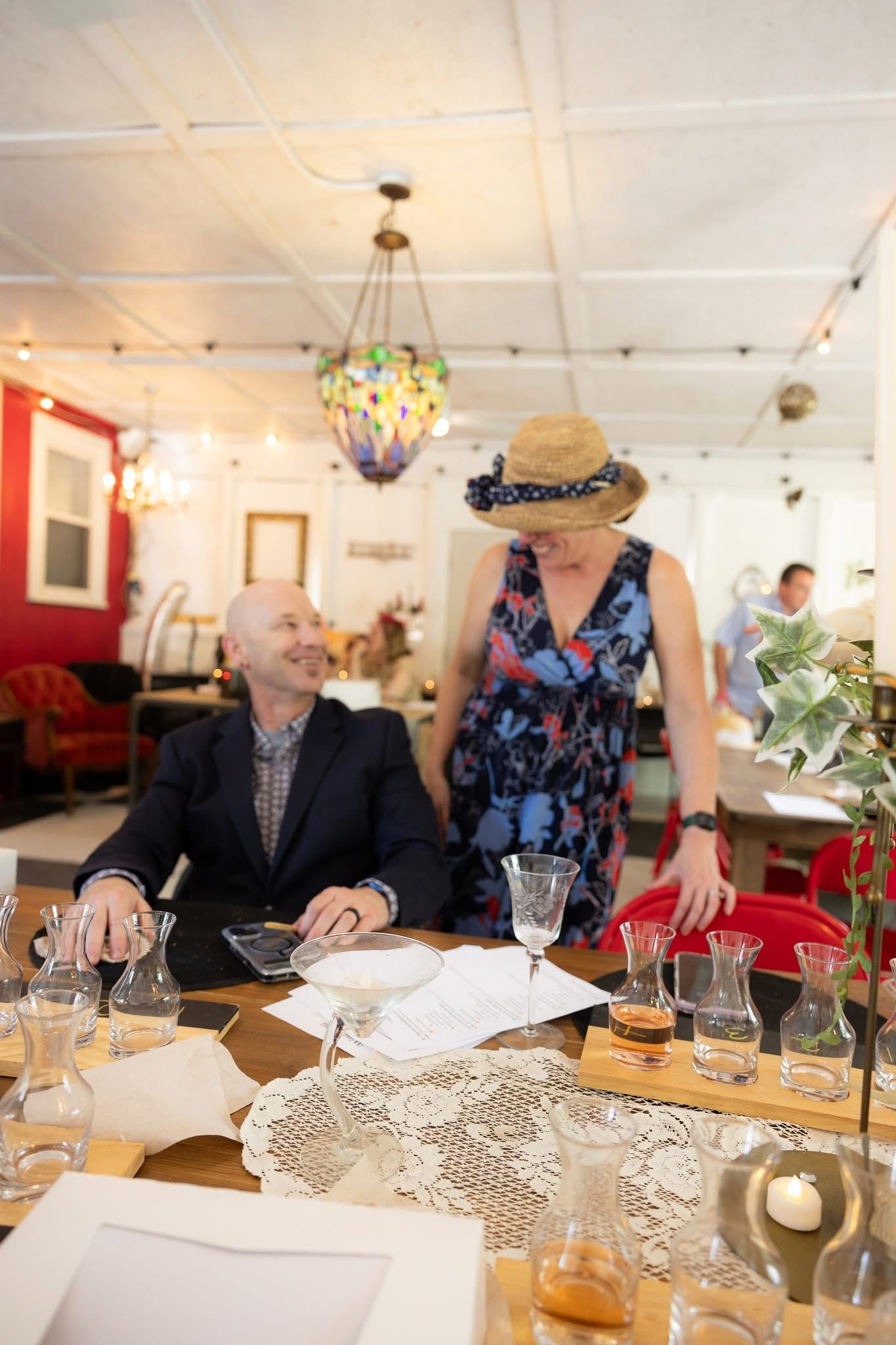 Man and woman at a table, smiling. She wears a hat, floral dress. Indoors, shop setting.