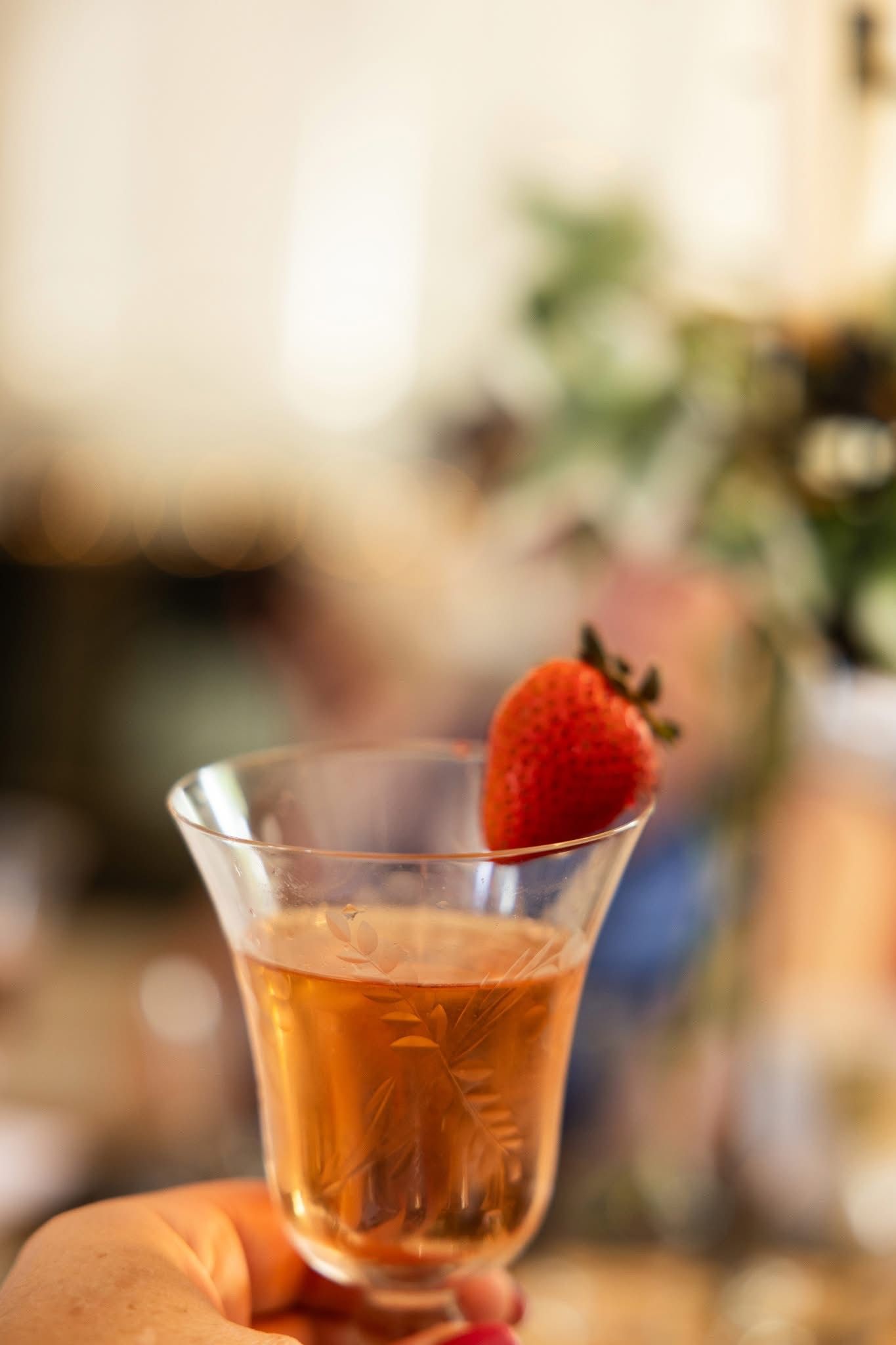 Hand holding a champagne glass with pink liquid, garnished with a strawberry. Blurred background.