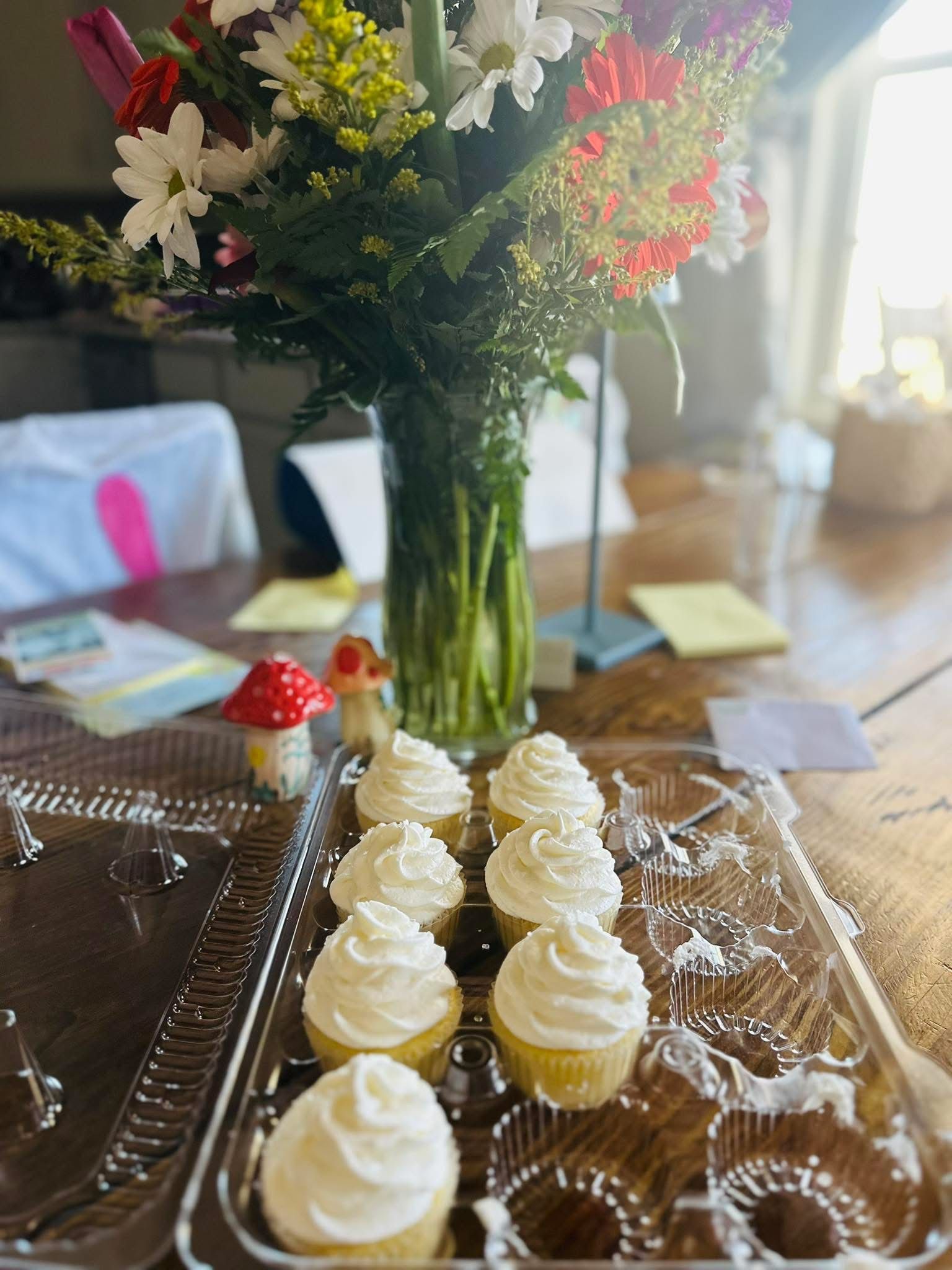 Cupcakes with white frosting in plastic container, flowers in vase on wooden table.