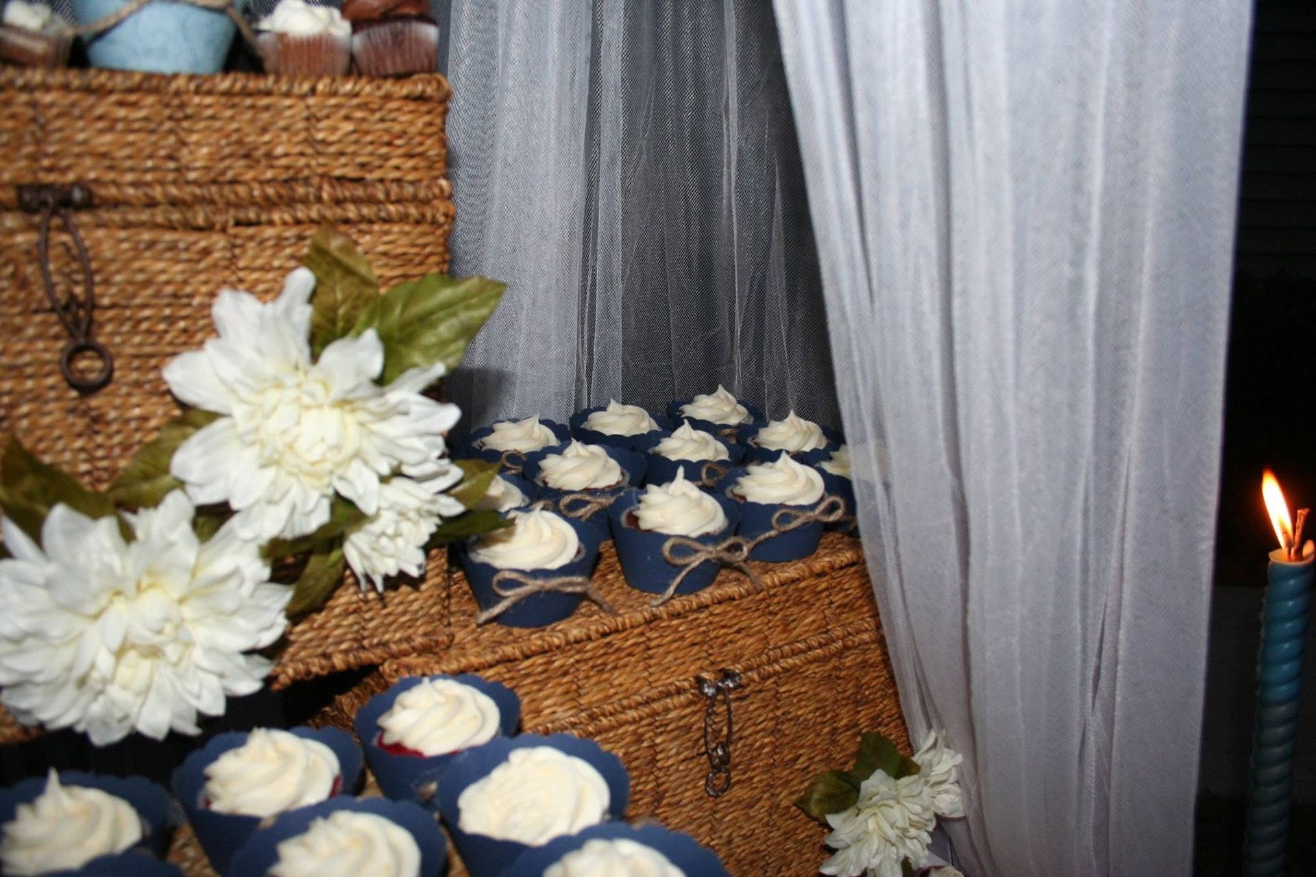 Cupcakes with white frosting and blue wrappers on woven baskets, white flowers, and draped white fabric.