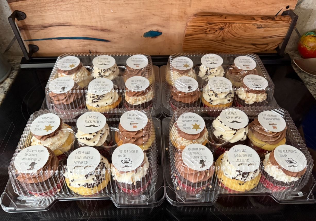 Cupcakes in clear containers, topped with labels. Various frosting and cake colors.