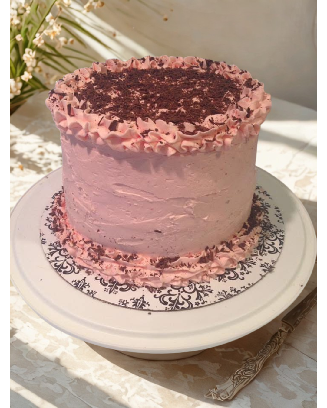 Pink frosted cake on a white cake stand, topped with chocolate shavings.
