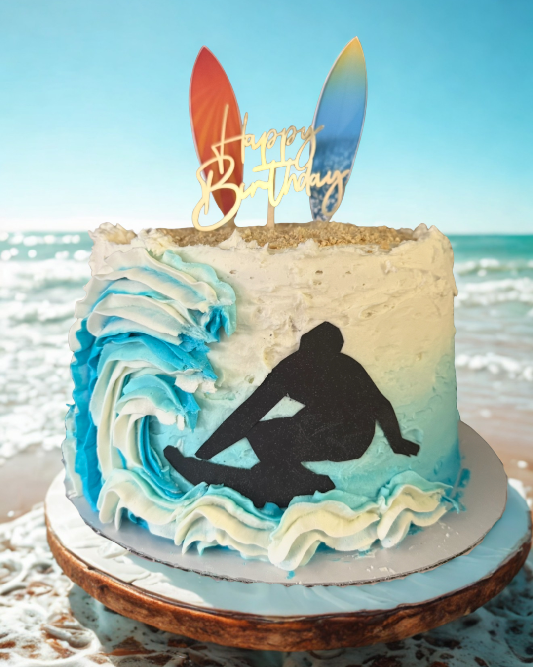 Birthday cake decorated with a surfer silhouette, wave, and surfboard toppers on a beach.