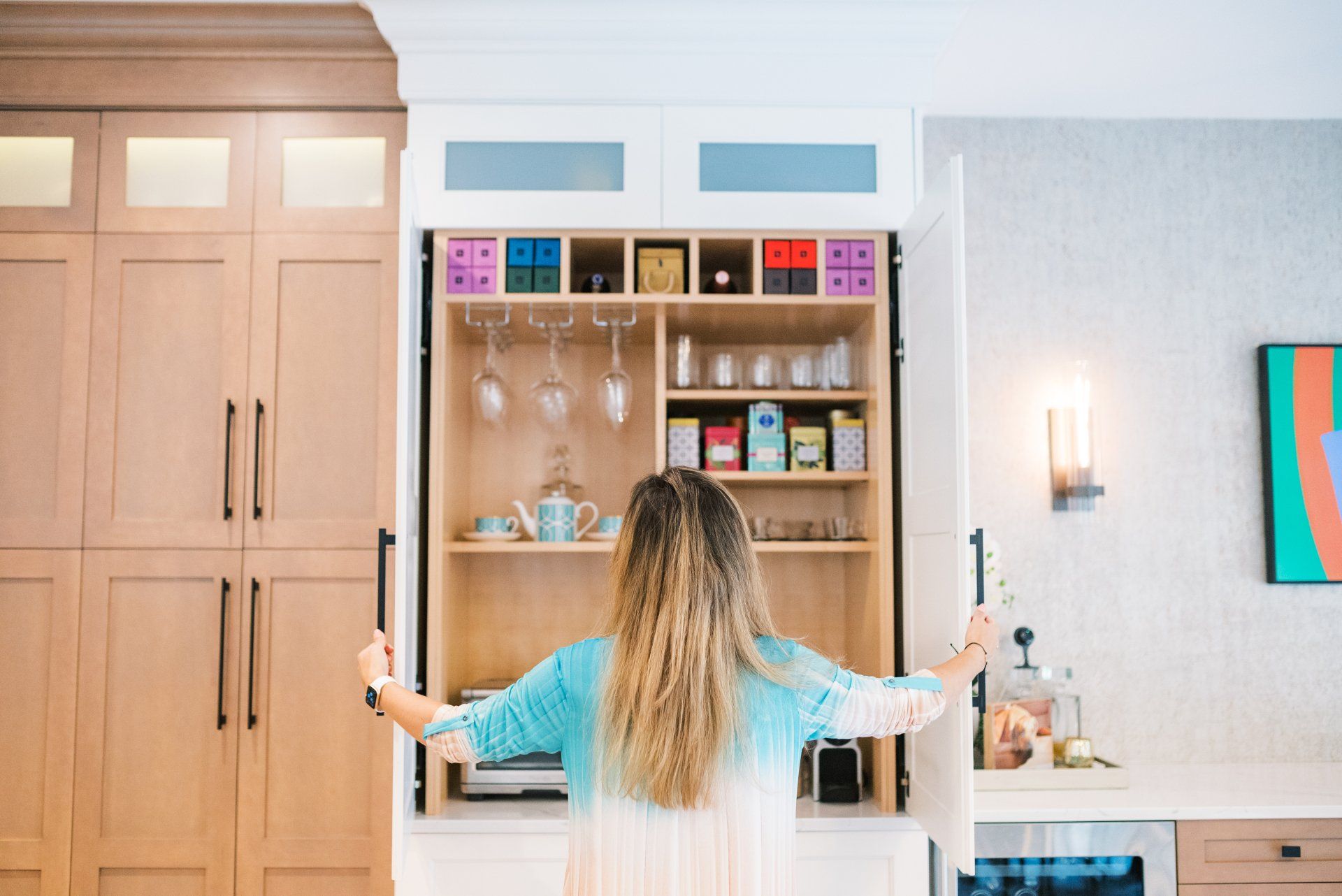 A woman is opening a cabinet in a kitchen with her arms outstretched.
