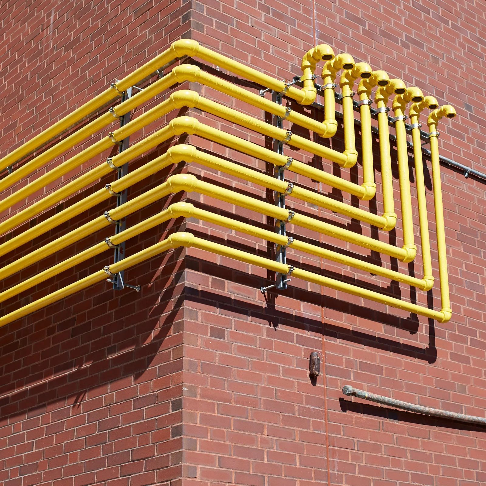 Yellow industrial pipes run along a red brick wall and turn upward at a corner.