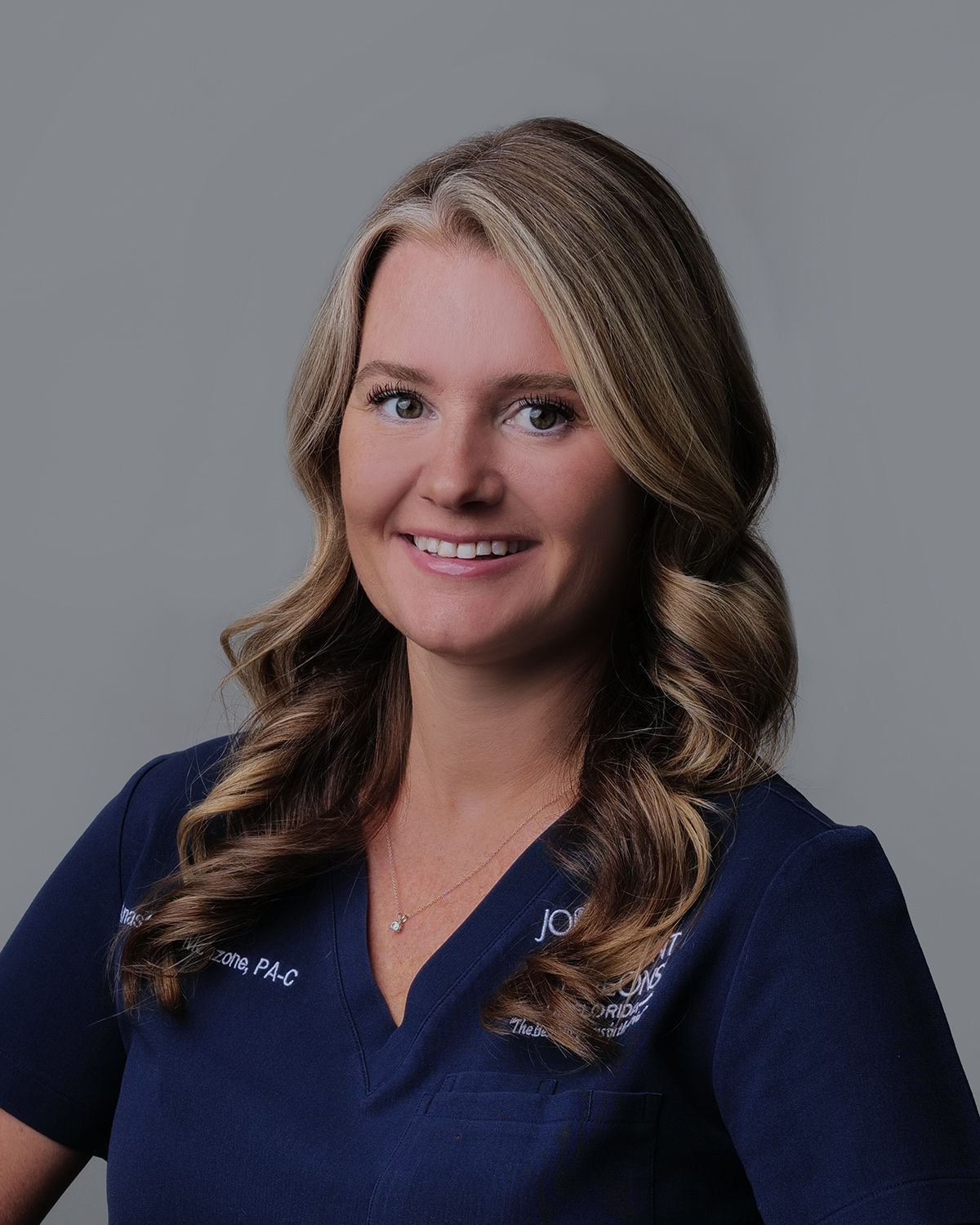 Woman with blonde hair in navy scrubs smiling, blurred surgical setting in background.
