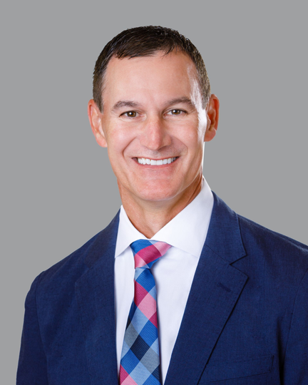 Smiling man in a blue suit and patterned tie, set against a blurred medical office background.
