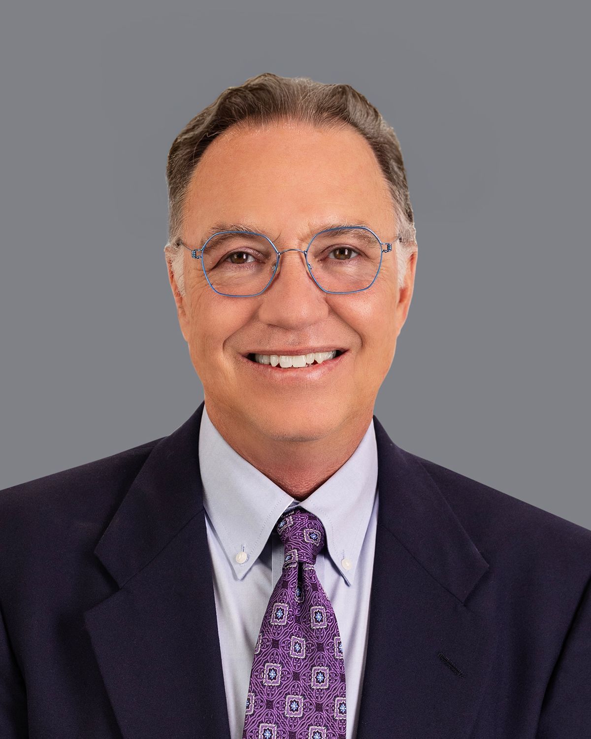 Smiling man in glasses, suit, and tie against a blurred medical setting.