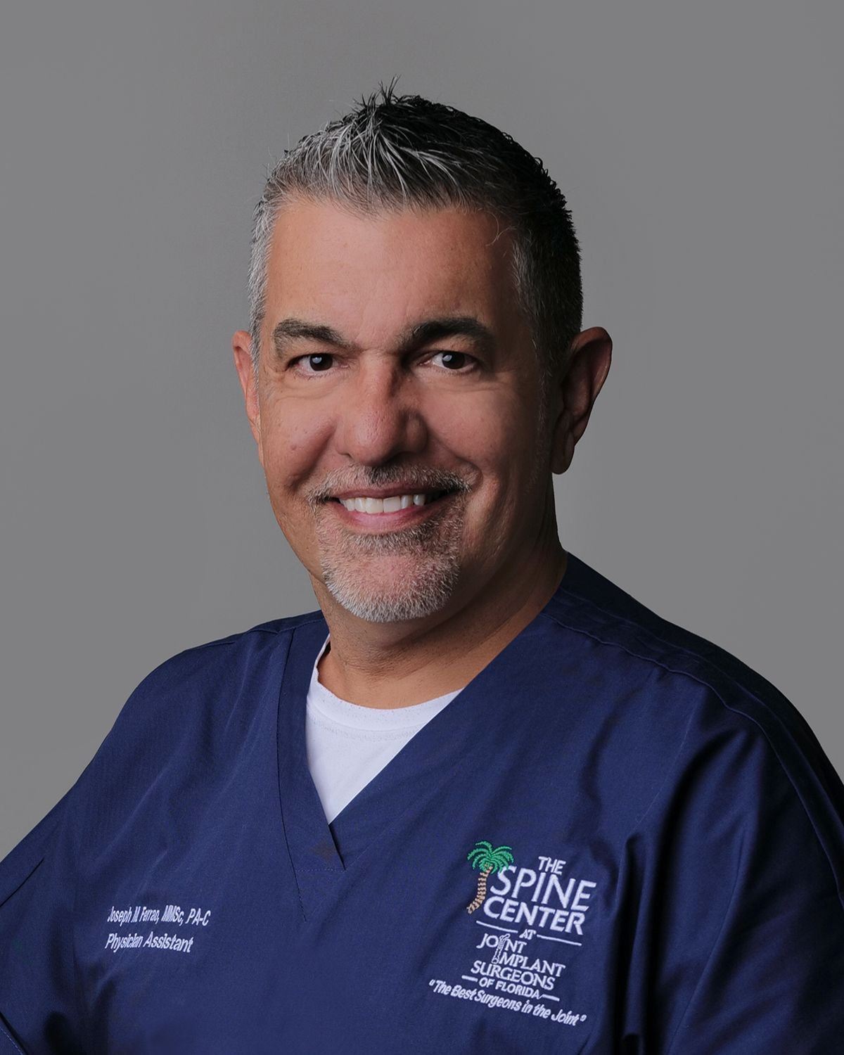 Man in navy suit with green tie, smiling against blurred hospital background.