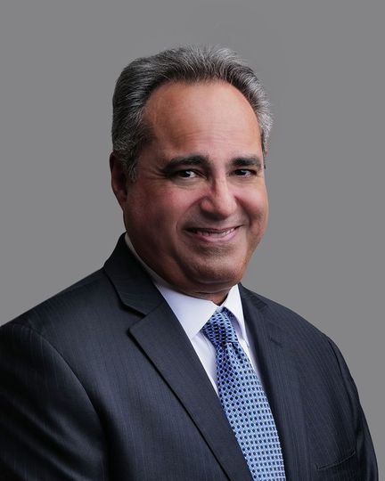 Smiling man in a suit and tie, possibly a doctor, in a blurred medical setting.