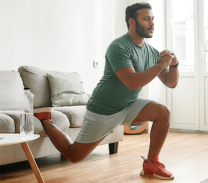 Man in athletic wear lunging in a living room, arms bent. Light wood floor, grey couch, water bottle.