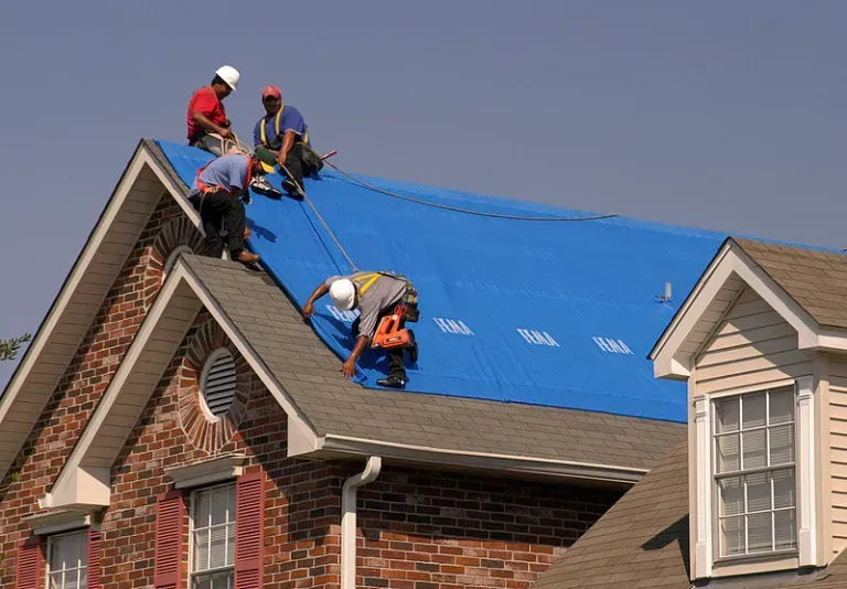 Workers on a house roof installing a blue tarp after storm damage