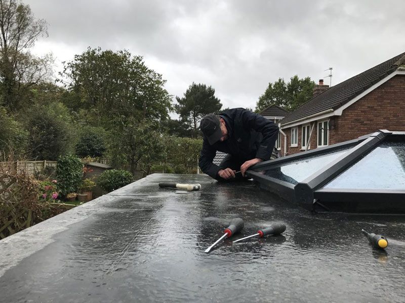 Worker repairing a flat black roof beside a skylight on a cloudy day