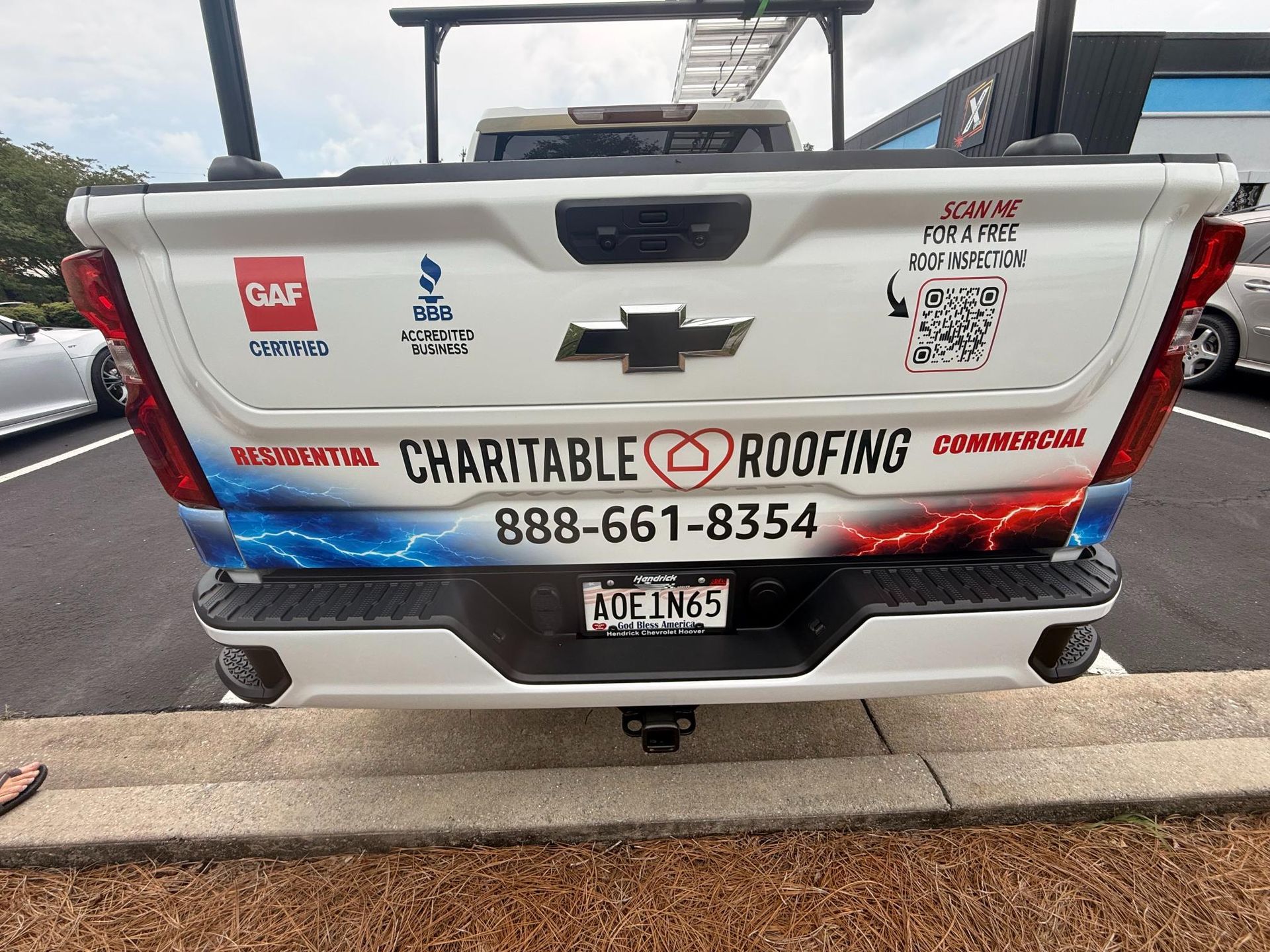 White Chevrolet pickup tailgate with Charitable Roofing advertising and phone number, parked in a lot