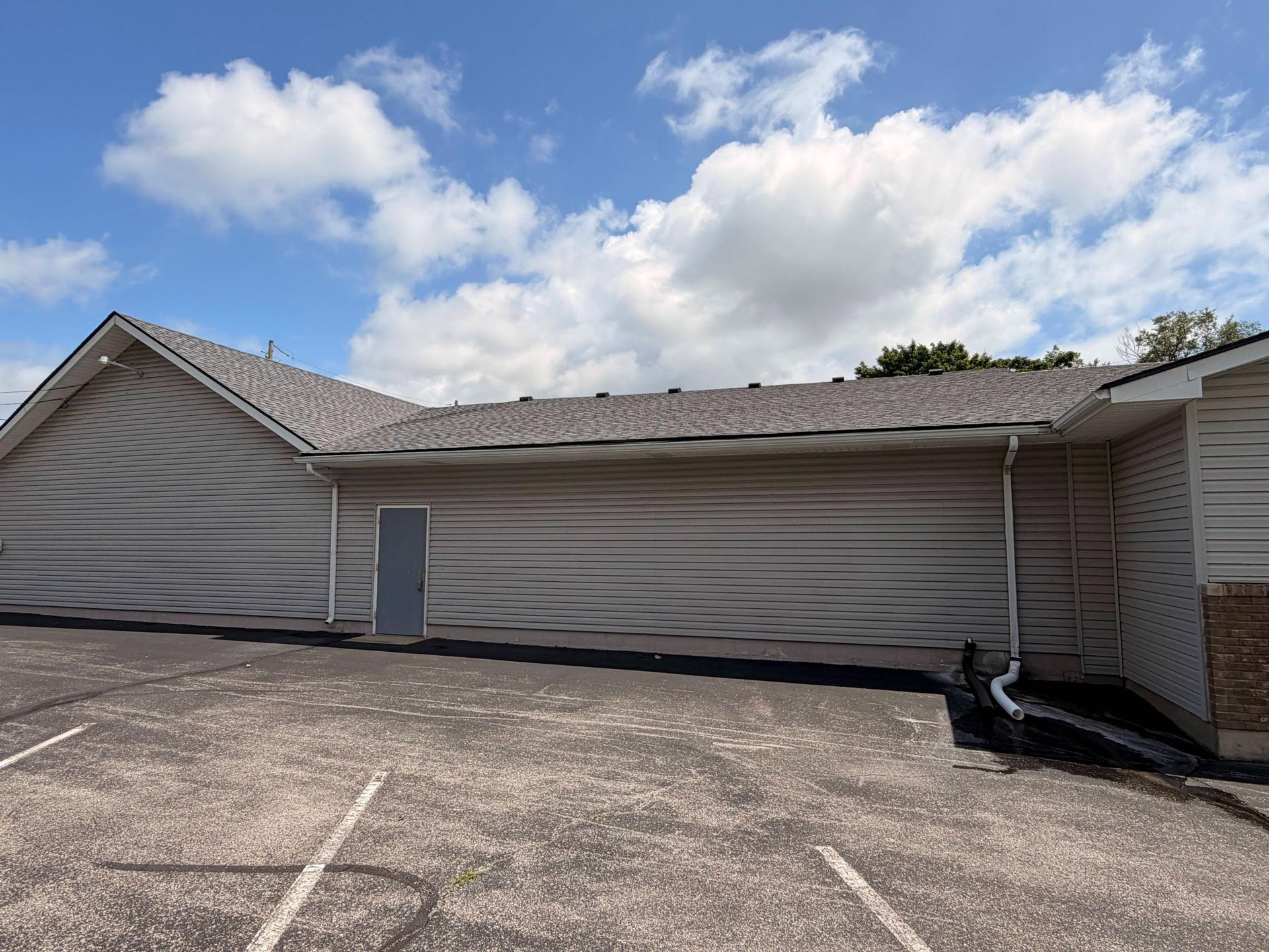 Low beige building with a gray roof and door, seen beside an empty parking lot under a blue sky.