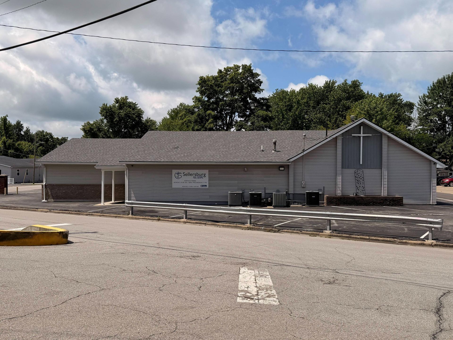 Small gray church building with a cross on the front, seen across a parking lot under a cloudy sky.