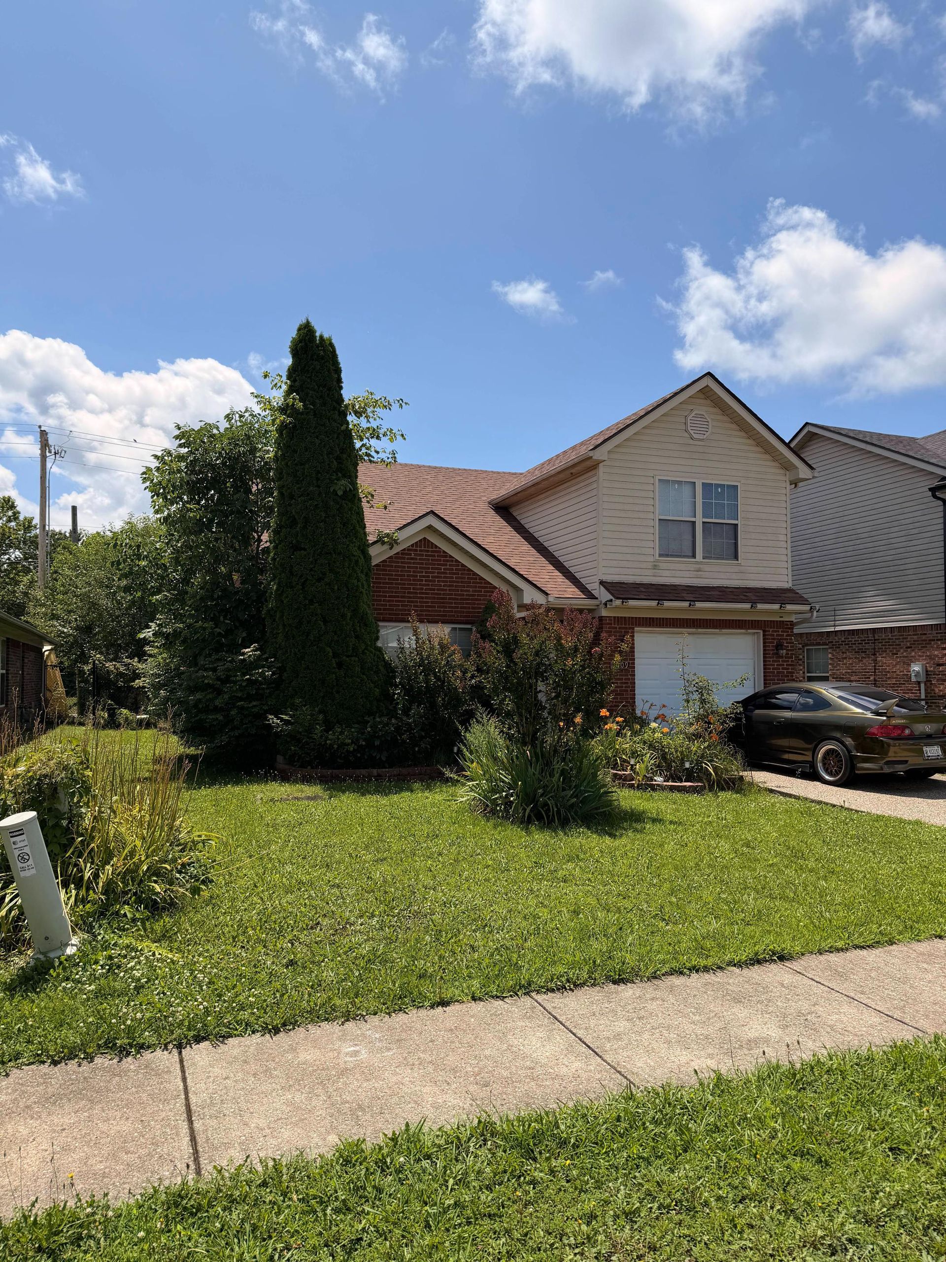 Suburban house with green lawn, tall tree, parked car, and partly cloudy blue sky