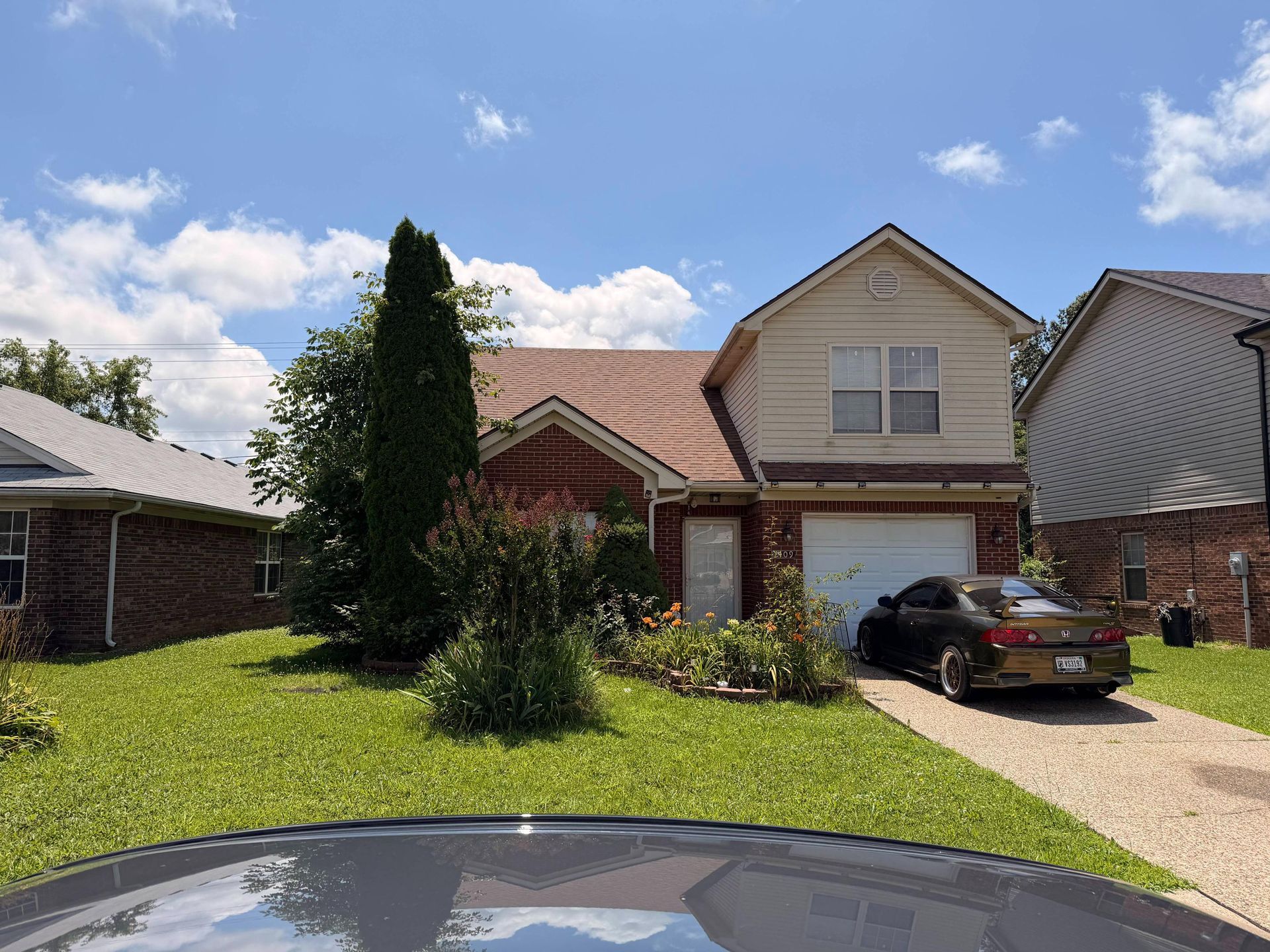 Suburban brick house with a front lawn, tall tree, and black car parked in the driveway.