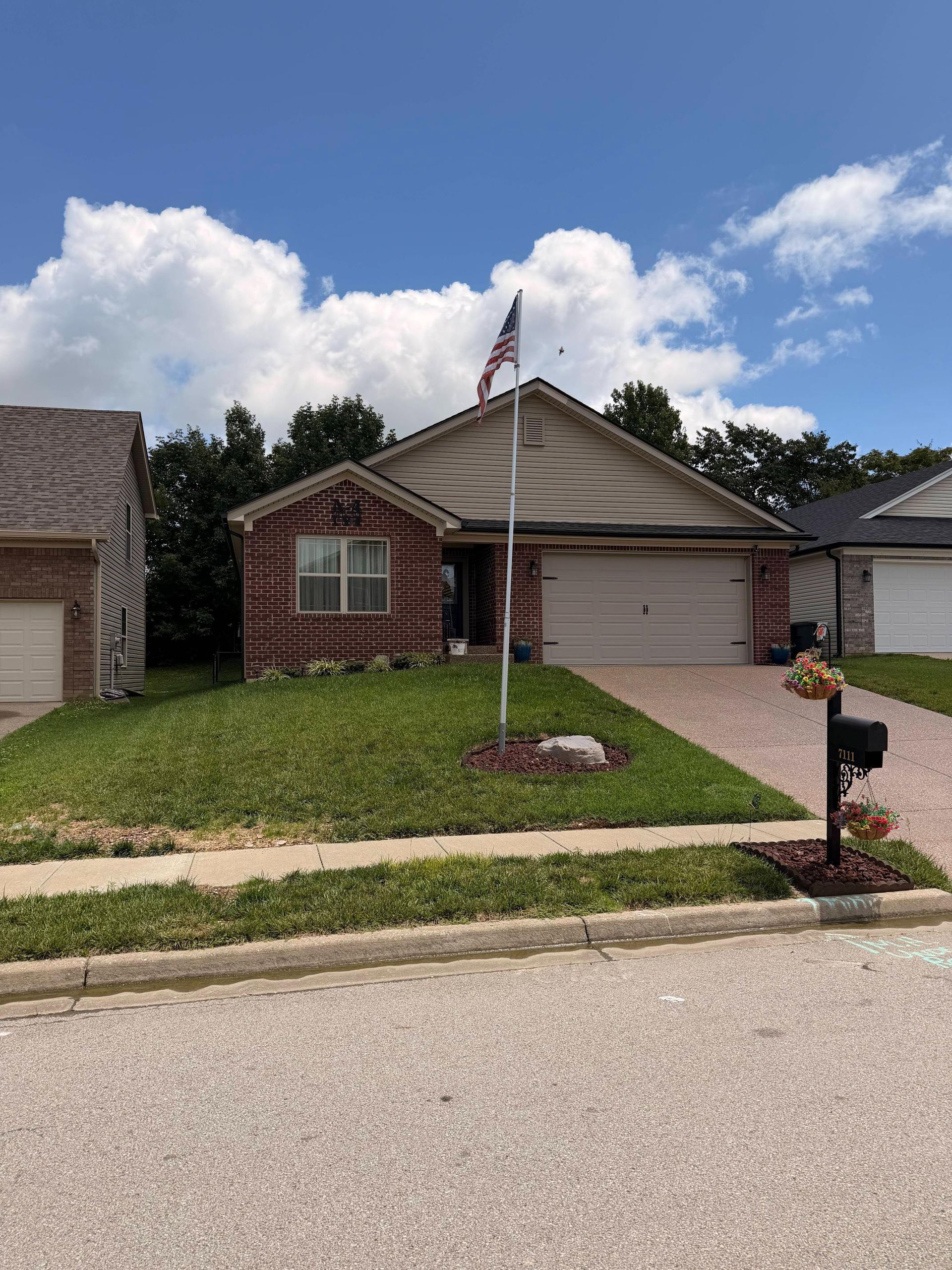 Suburban brick house with a front lawn, garage, flagpole, and mailbox under a partly cloudy sky