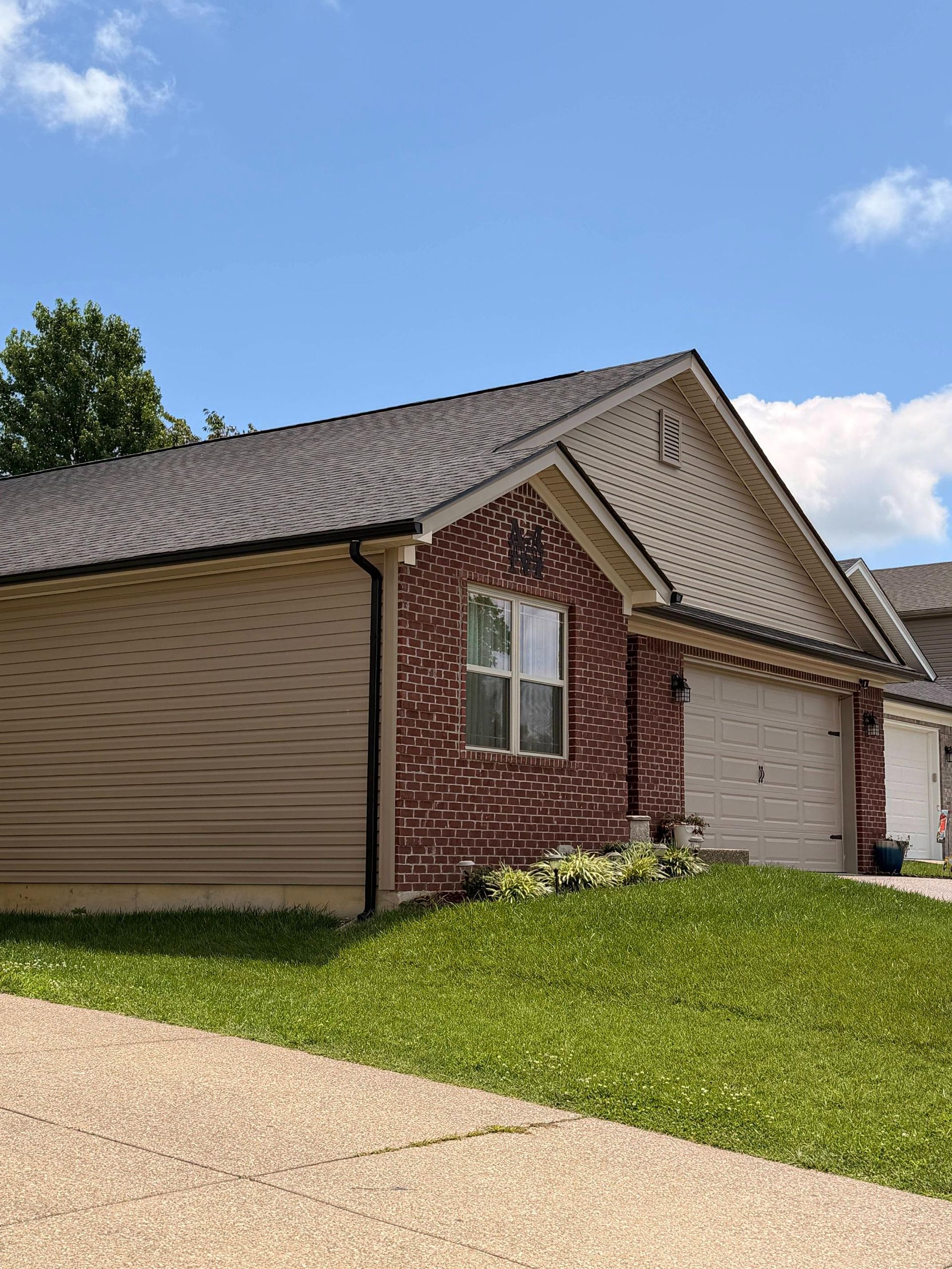 Suburban brick-and-beige house with garage, sloped roof, and green lawn under a blue sky
