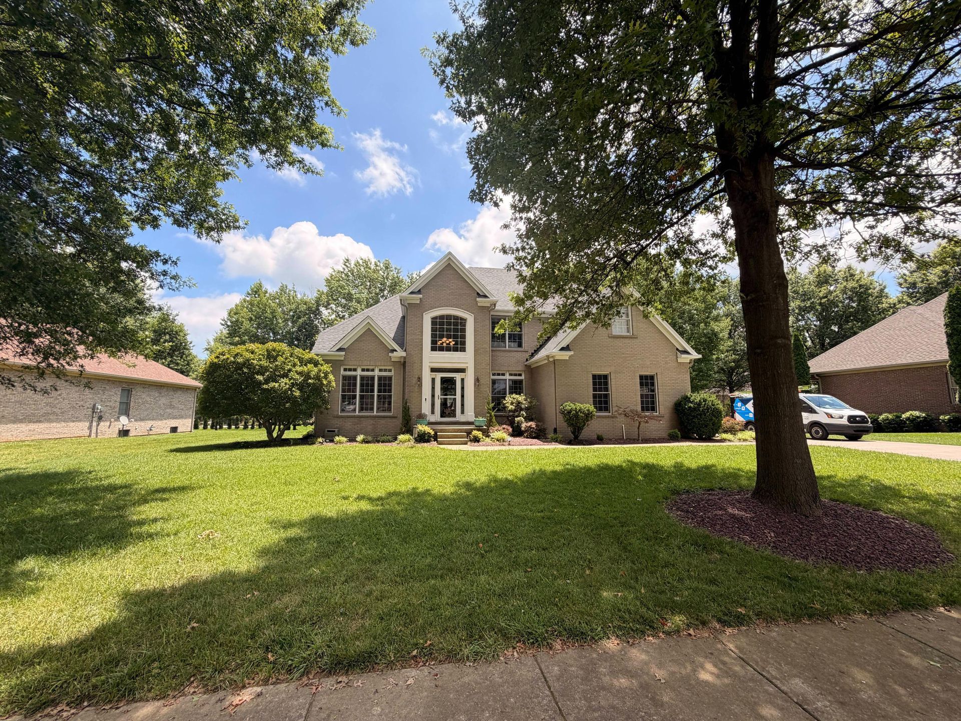 Large beige suburban house with a front lawn, framed by trees under a blue sky