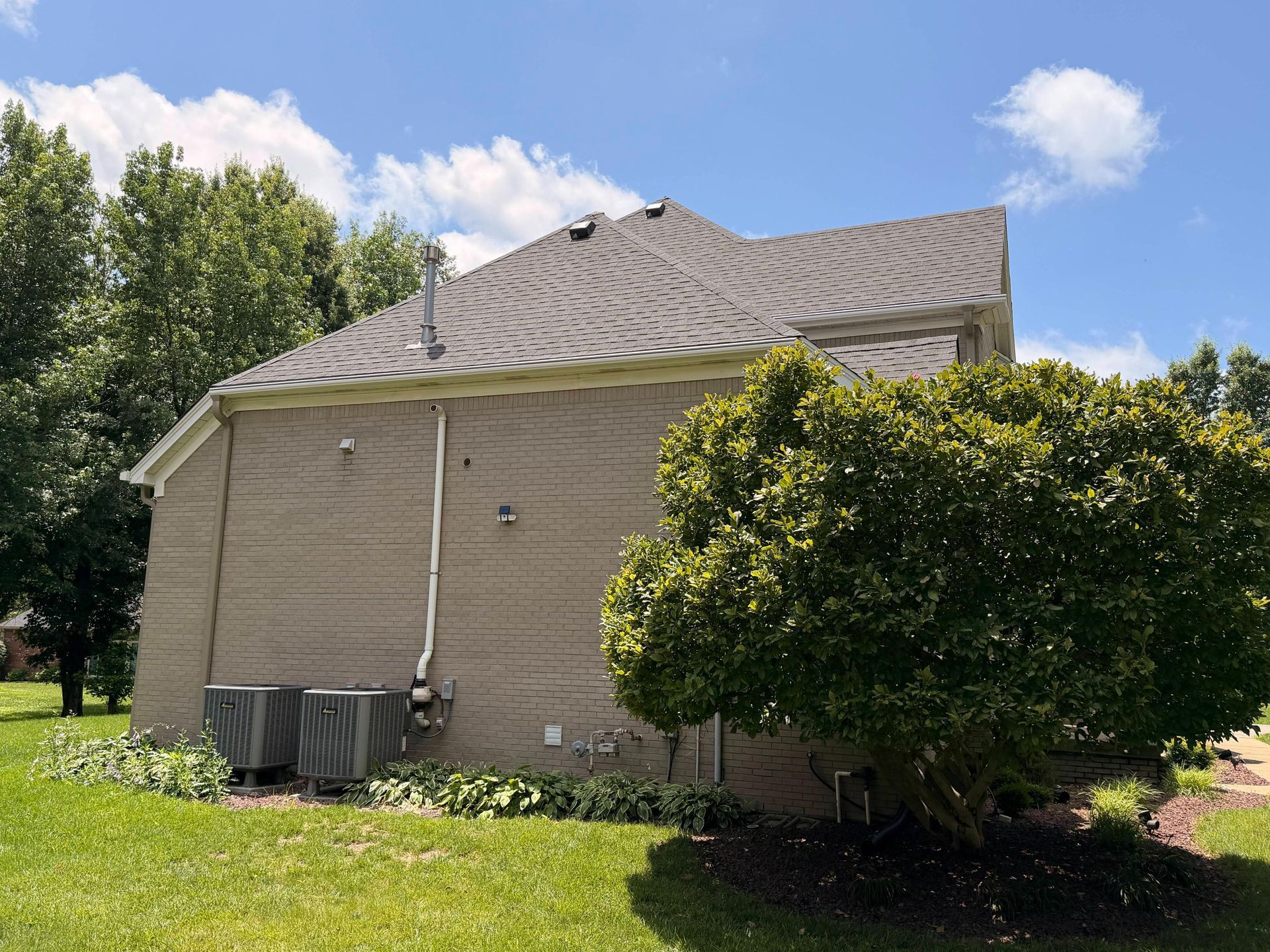Side view of a beige house with a shingled roof, bushes, and air-conditioning units in a grassy yard under a blue sky