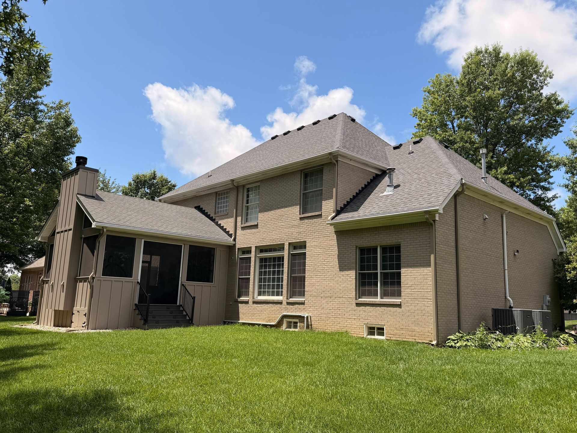 Tan brick house with a gray roof, covered porch, and green lawn under a blue sky